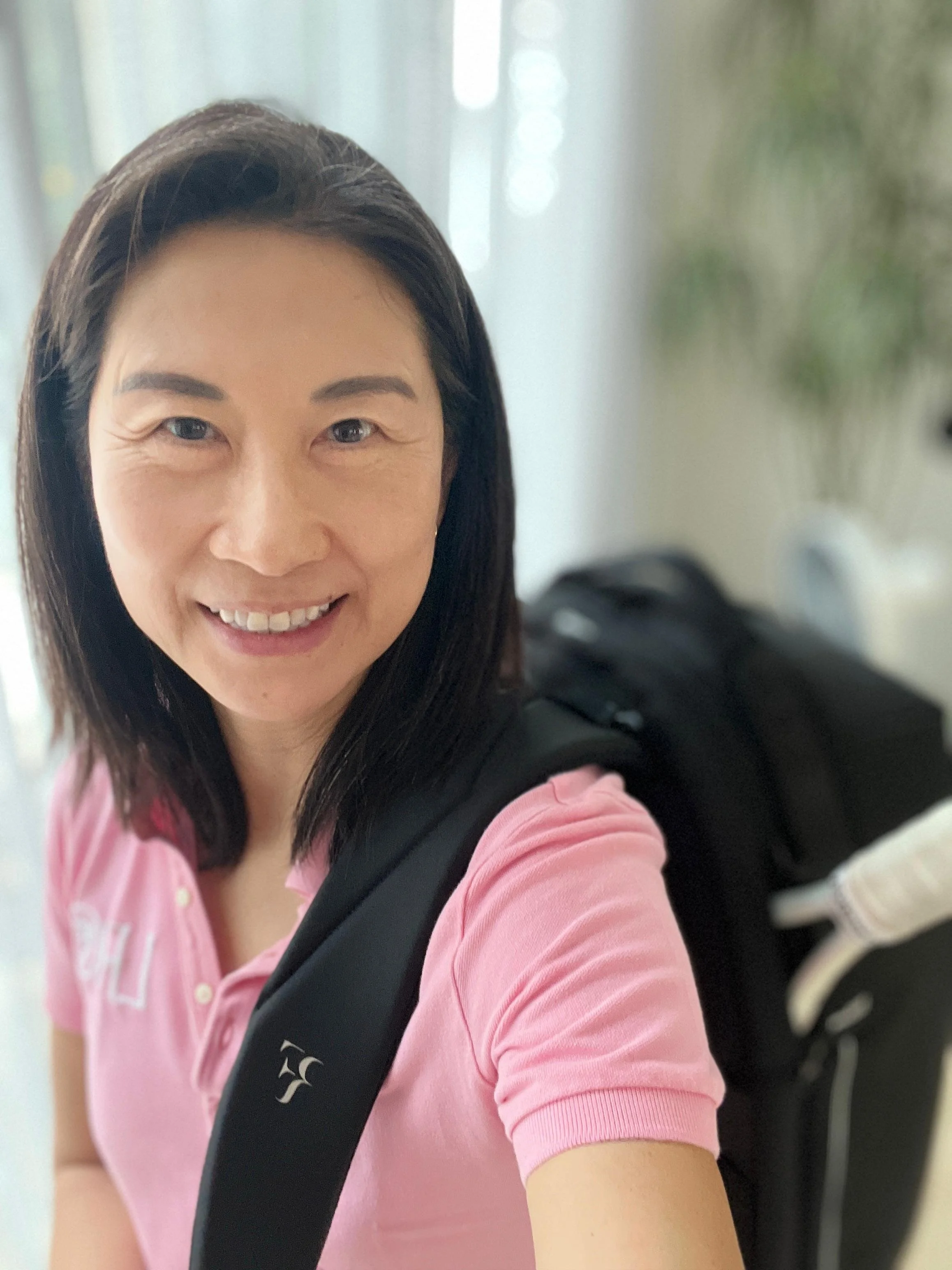 A woman with shoulder-length dark hair taking a selfie, smiling, wearing a pink polo shirt and carrying a black backpack, with a blurred background of windows and indoor plants.