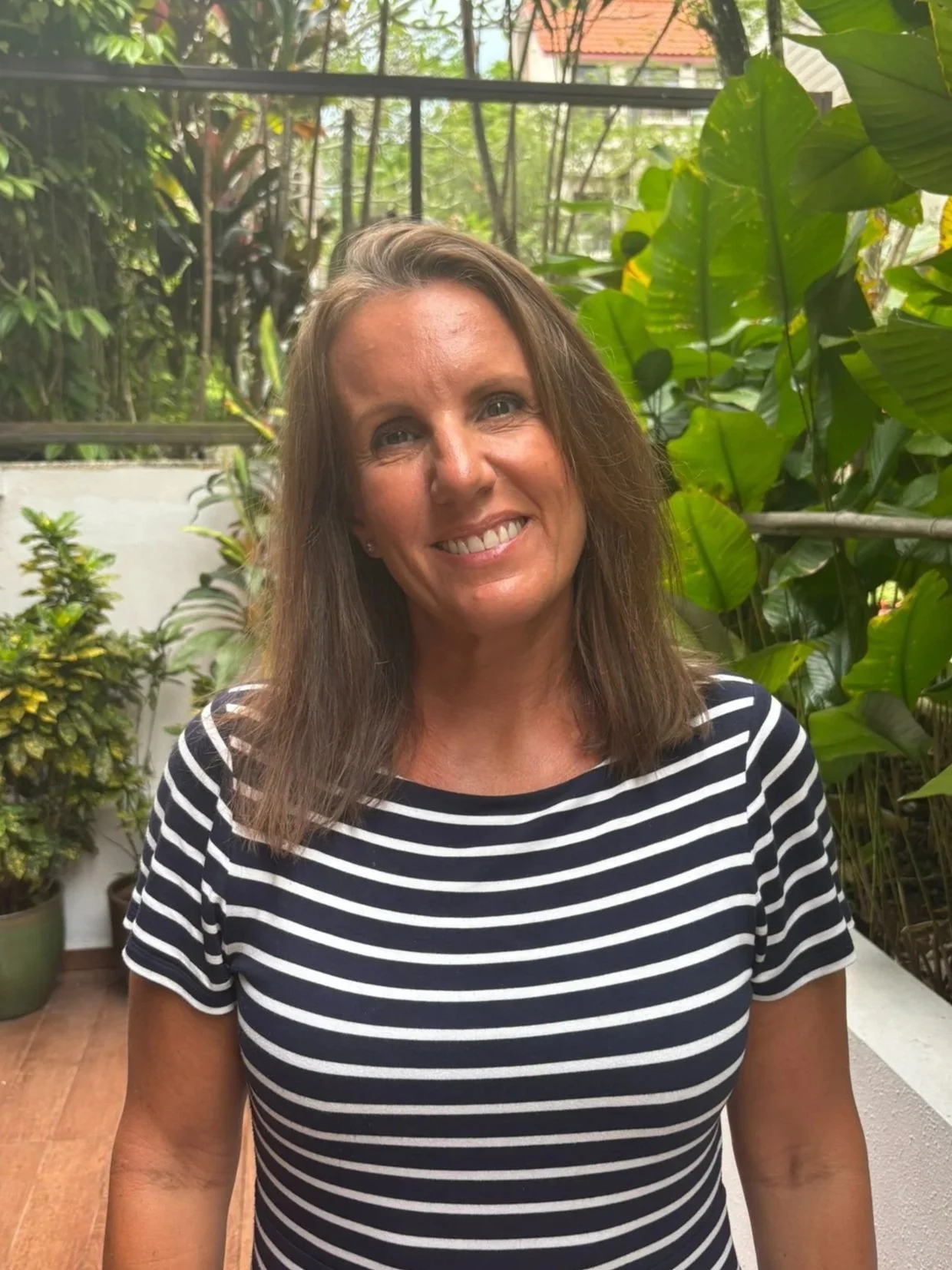 A smiling woman with shoulder-length brown hair wearing a black and white striped shirt standing in a lush green indoor garden with various plants and a glass window in the background.