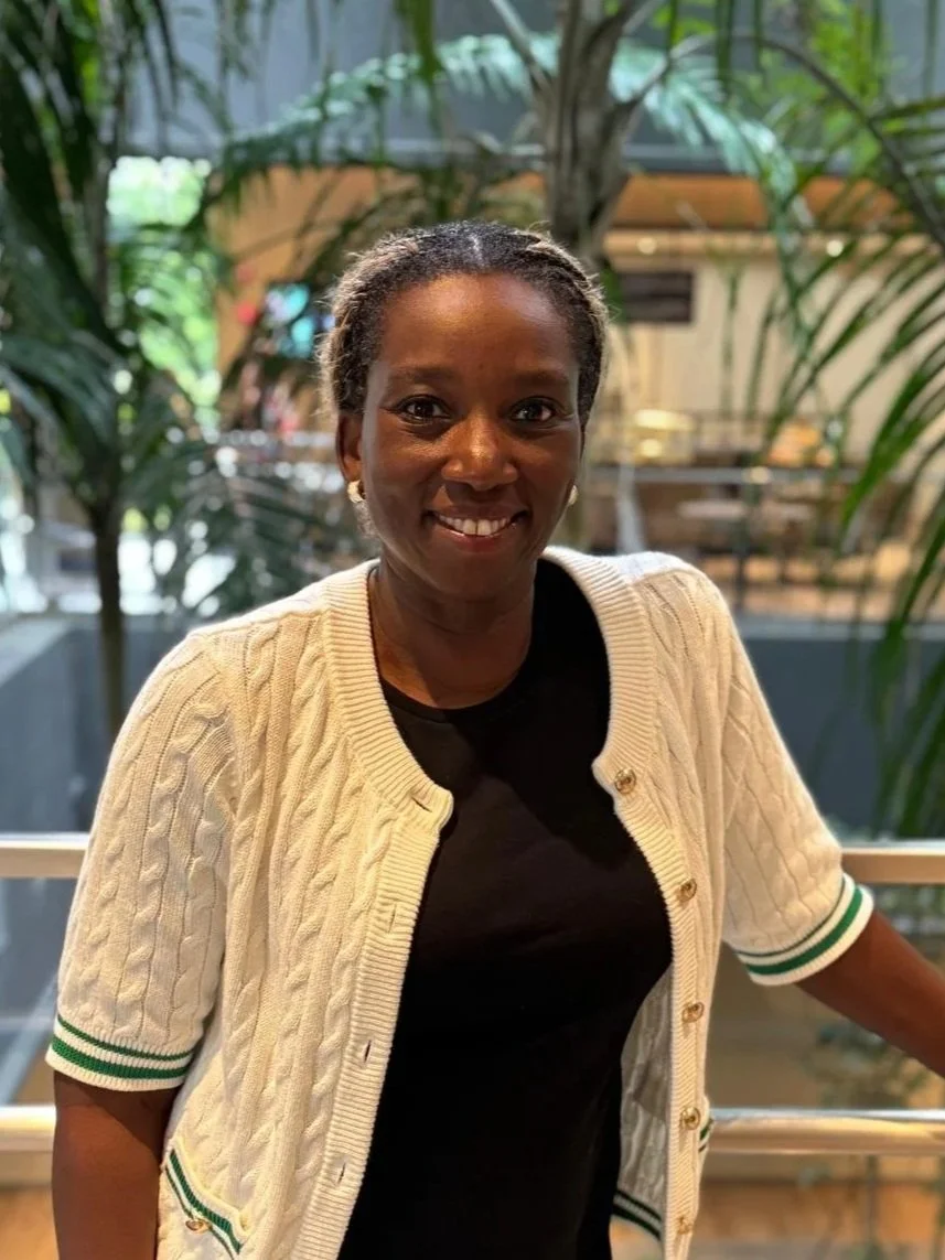 A smiling woman with short, curly hair standing indoors in front of greenery and a blurred background, wearing a beige cardigan with green and white striped cuffs over a black shirt.