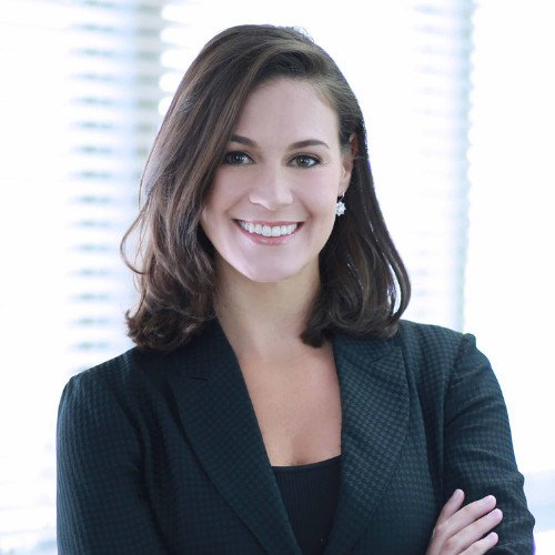 A smiling woman with short, curly hair standing indoors in front of greenery and a blurred background, wearing a beige cardigan with green and white striped cuffs over a black shirt.