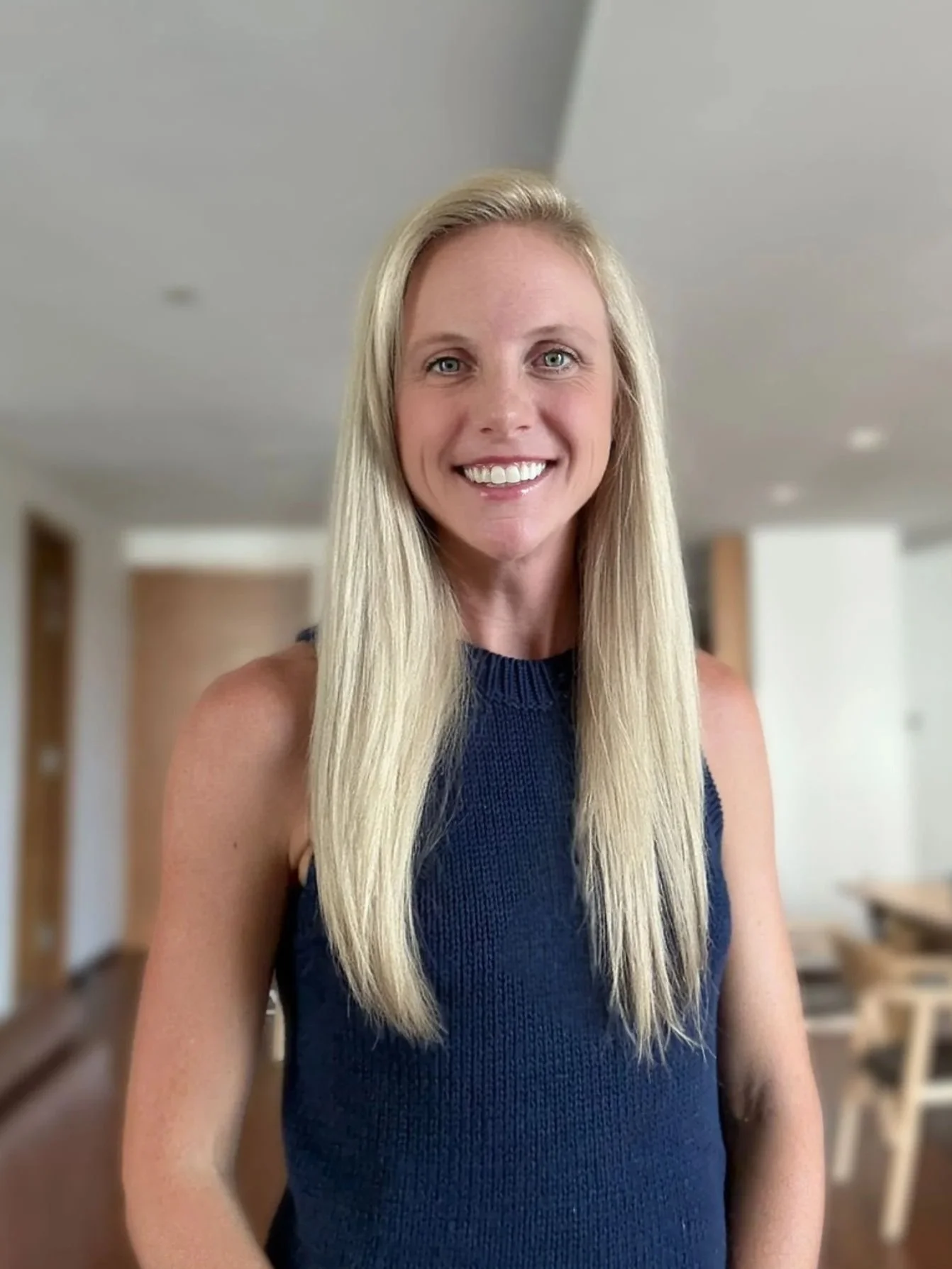 A smiling blonde woman in a navy sleeveless top standing indoors in front of a blurred background with wooden furniture.