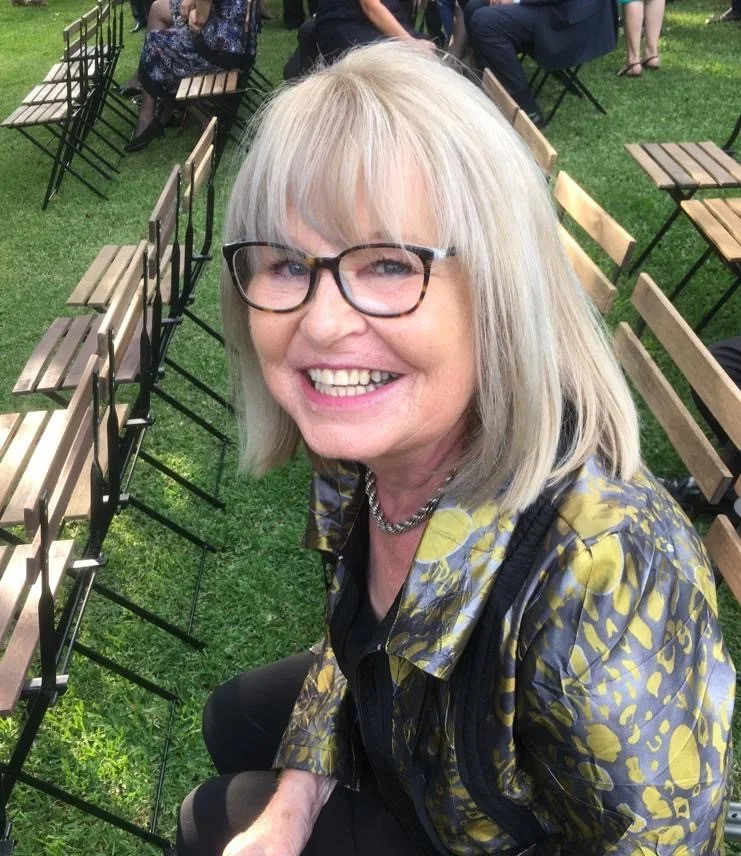 A smiling older woman with gray hair, glasses, and a patterned jacket sitting outdoors on grass with empty chairs around her.