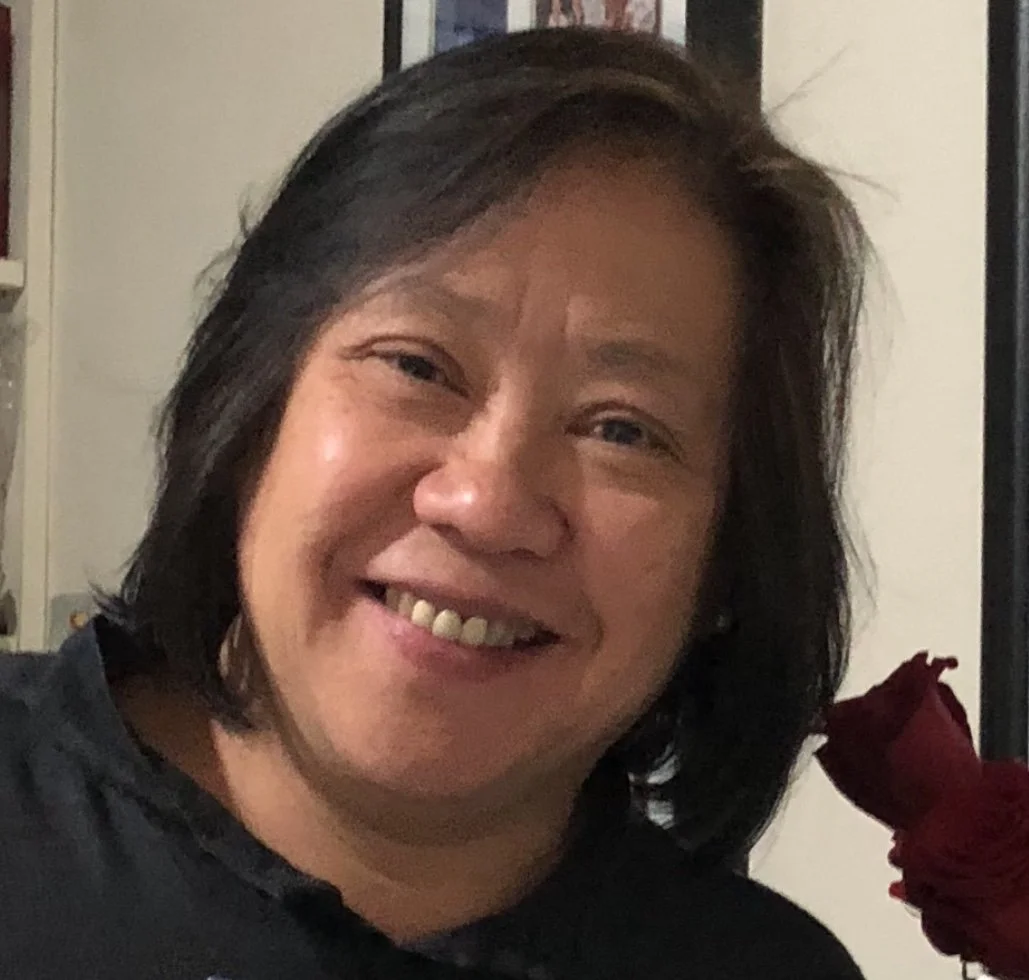 A woman with medium-length black hair smiling at the camera, wearing a black top, with a red rose partially visible on the right side of the image.