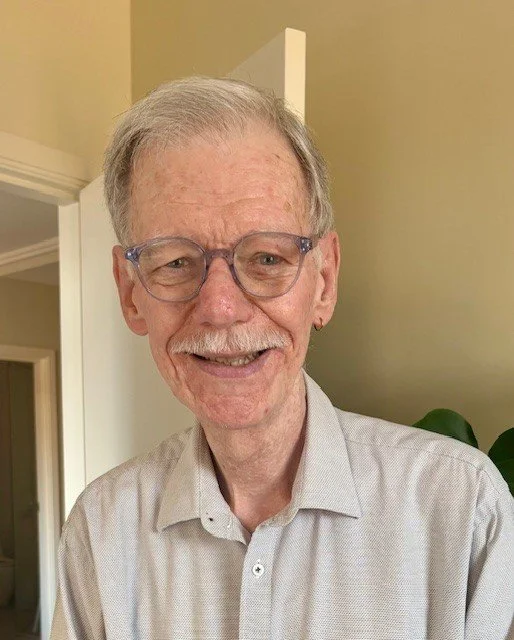 Smiling elderly man with glasses and a light collared shirt indoors.