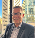 A man with glasses wearing a suit jacket and collared shirt, standing indoors with tall buildings visible through the window behind him.