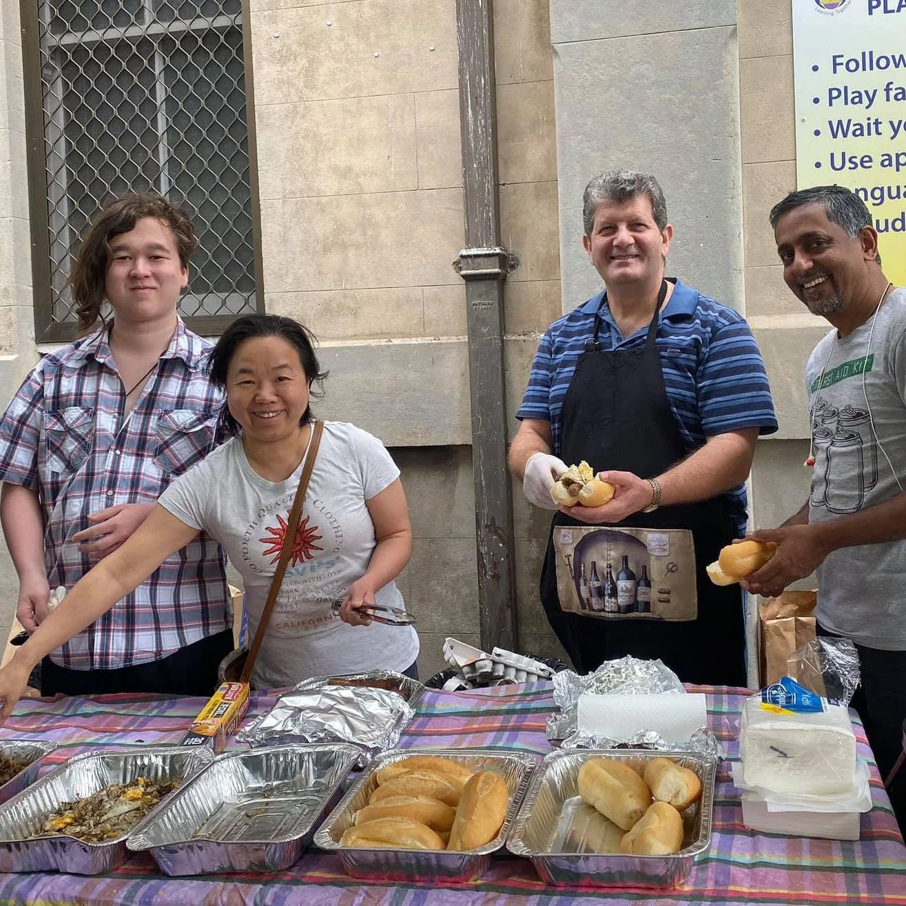 Four people standing behind a table with hot dog buns, foil trays, and food at a street food stall, smiling and holding hot dogs.