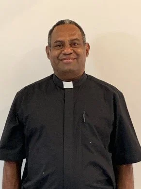 A smiling man in a black clergy shirt standing against a plain wall.