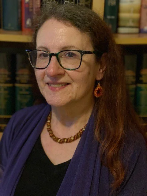 A woman with long reddish-brown hair, glasses, and earrings, smiling, standing in front of a bookshelf filled with books.