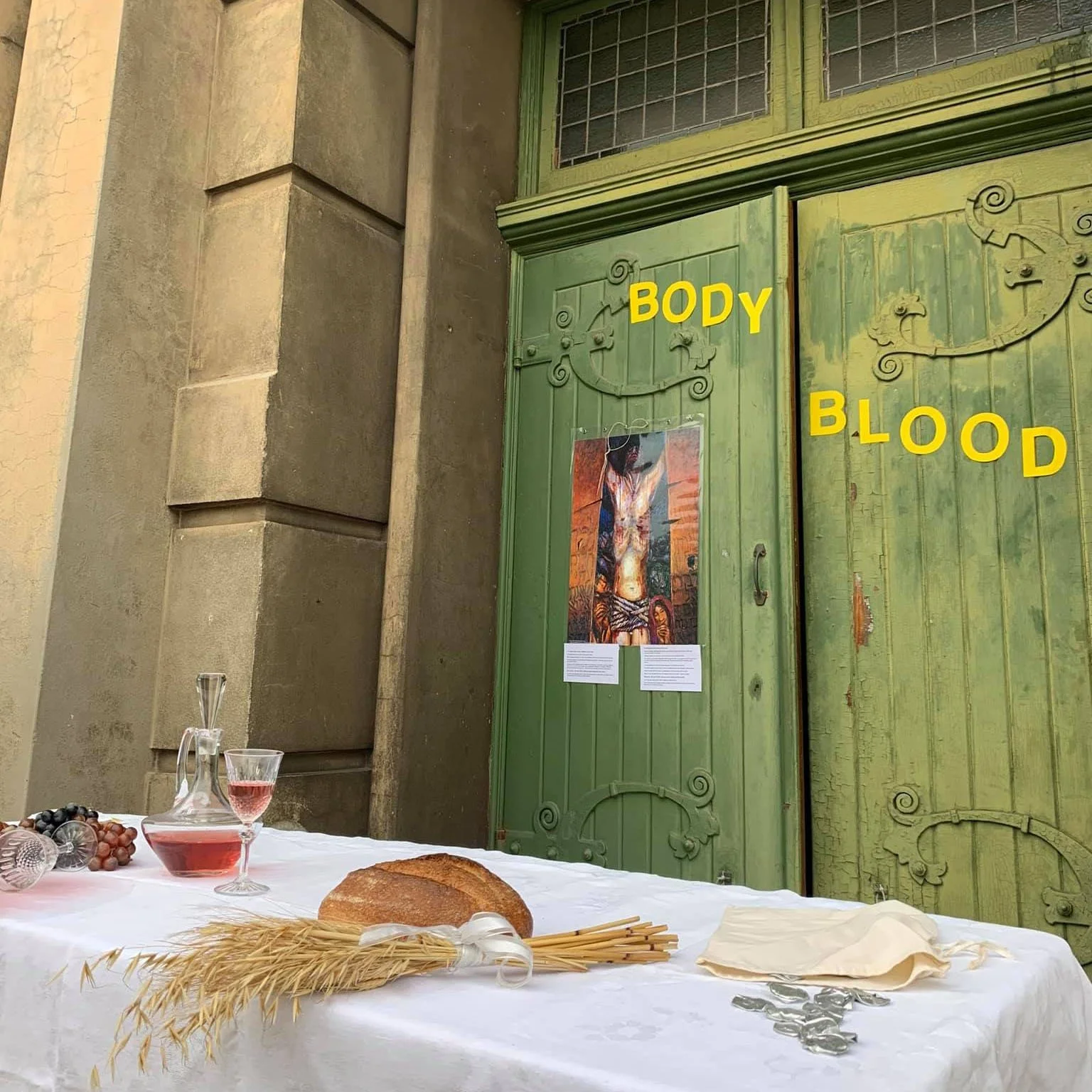 A table with bread, grapes, a decanter of red wine, a filled wine glass, and silver jar covers in front of a green door with the words 'BODY' and 'BLOOD' in yellow letters, and a religious art piece on the door.