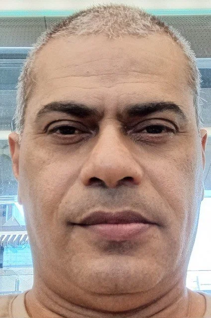 Close-up of a man with short gray hair and light skin, looking at the camera indoors with a window and building reflections in the background.