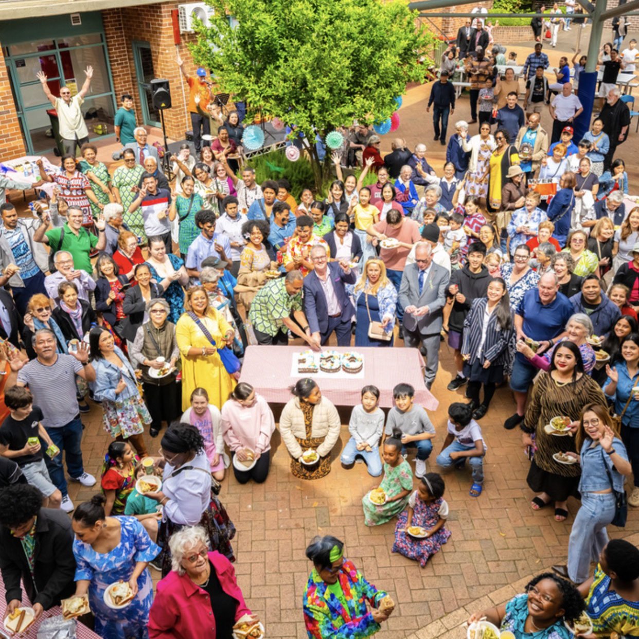 A large, diverse group of people celebrating outdoors around a decorated table with cakes, with children in traditional clothing and many smiling and waving.