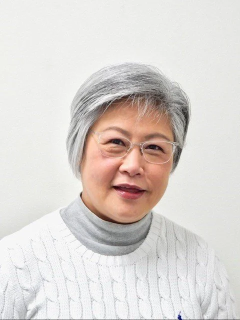A middle-aged woman with short gray hair wearing glasses, a light gray turtleneck, and a white cable-knit sweater, smiling at the camera against a plain white background.