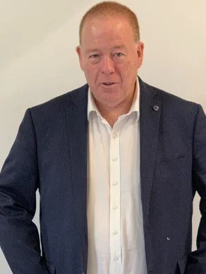 A middle-aged man with short red hair, wearing a dark blue blazer over a white shirt, standing against a plain light-colored wall.