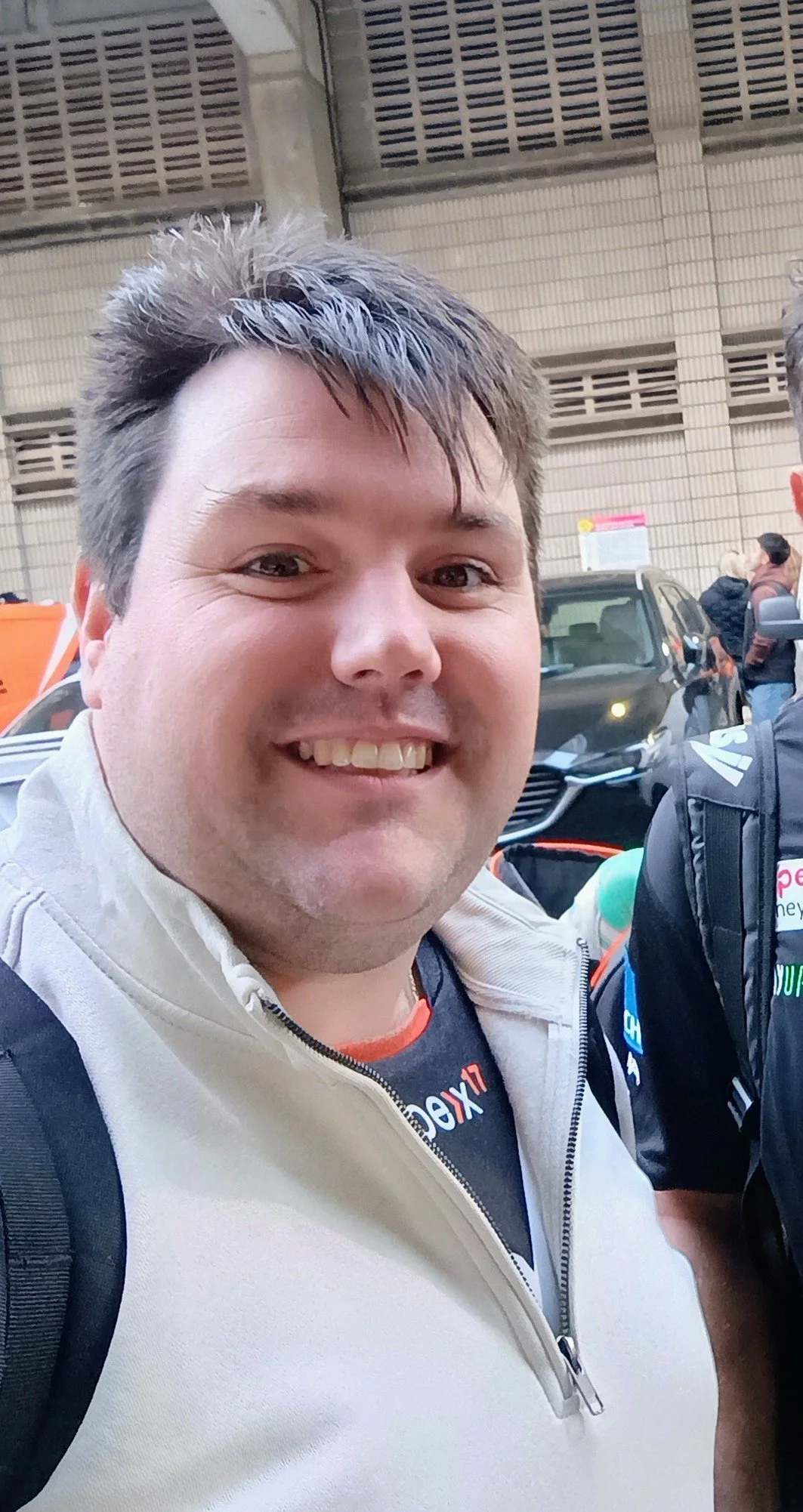 Close-up of smiling man at a busy outdoor event with people and vehicles in the background.