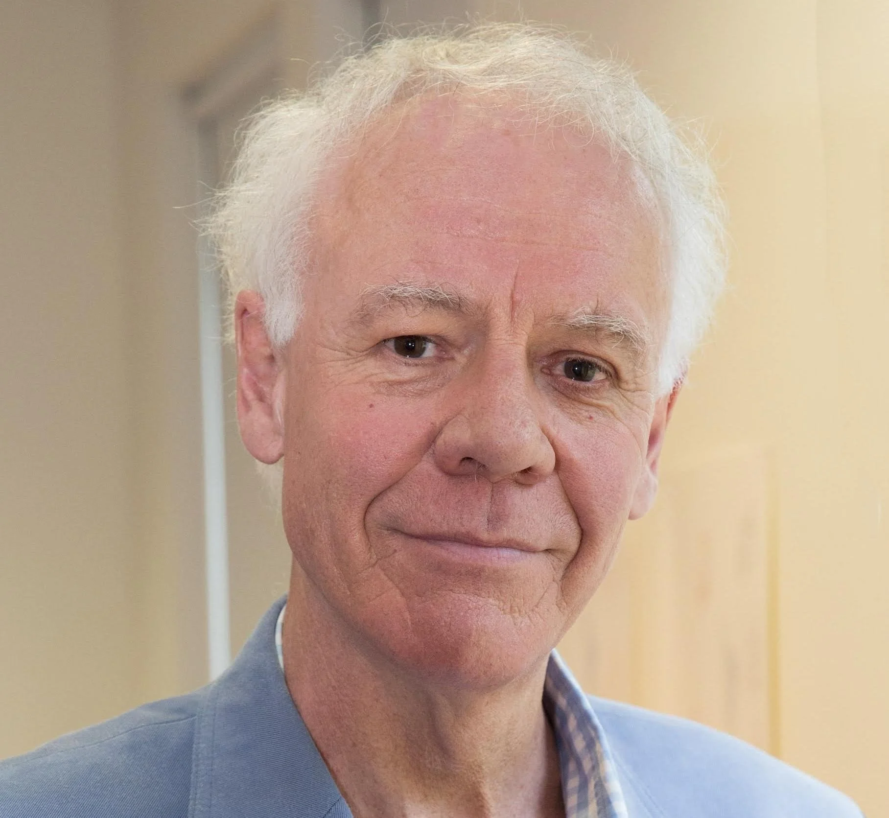 Portrait of an elderly man with white hair, wearing a blue blazer and a checkered shirt, smiling indoors.