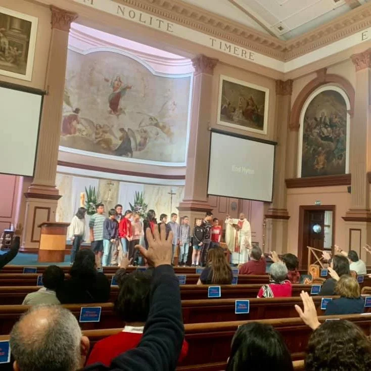 Group of children on stage in a church or cathedral, with a priest or pastor, congregation seated in pews, some raising hands, large religious paintings and screens in the background.