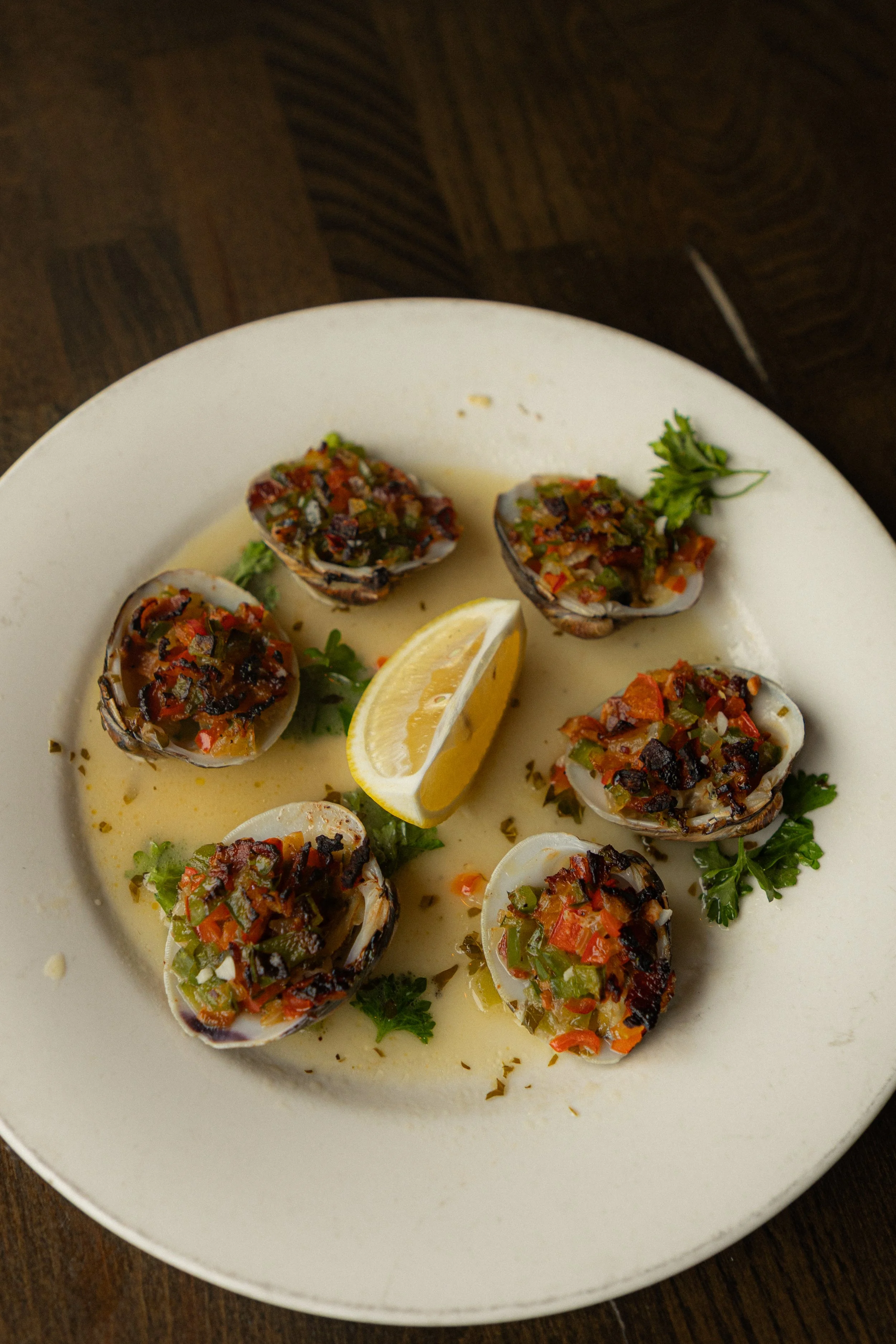 On a white round plate, 6 clams filled with a blend of bacon, breading, & peppers. In the middle a lemon wedge