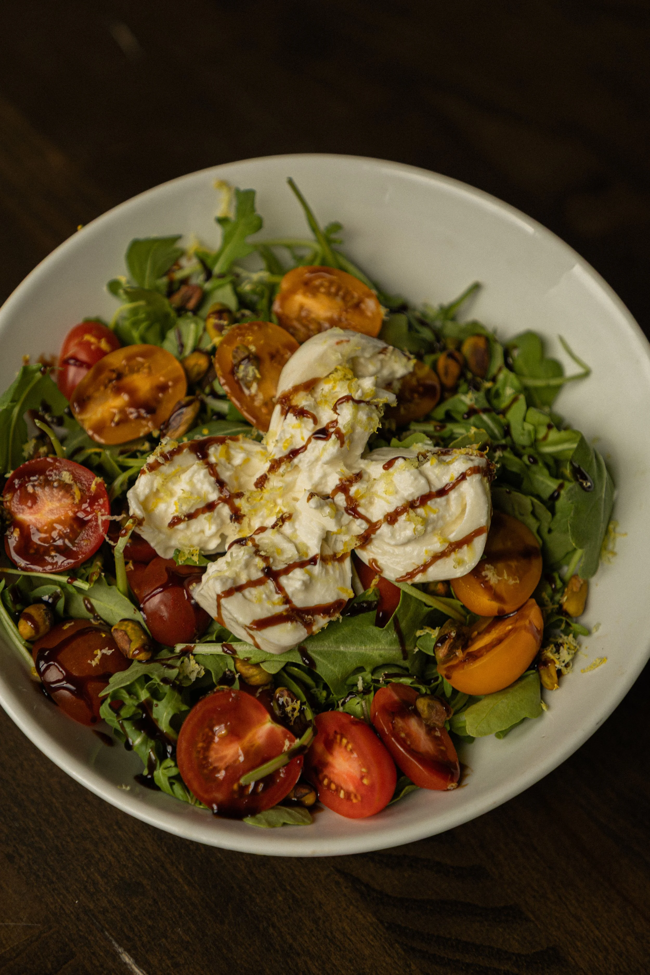 Sliced open Burrata tops a bowl of arugula, with cherry tomatoes, pistachios mixed in. Drizzled with balsamic glaze & lemon zest..