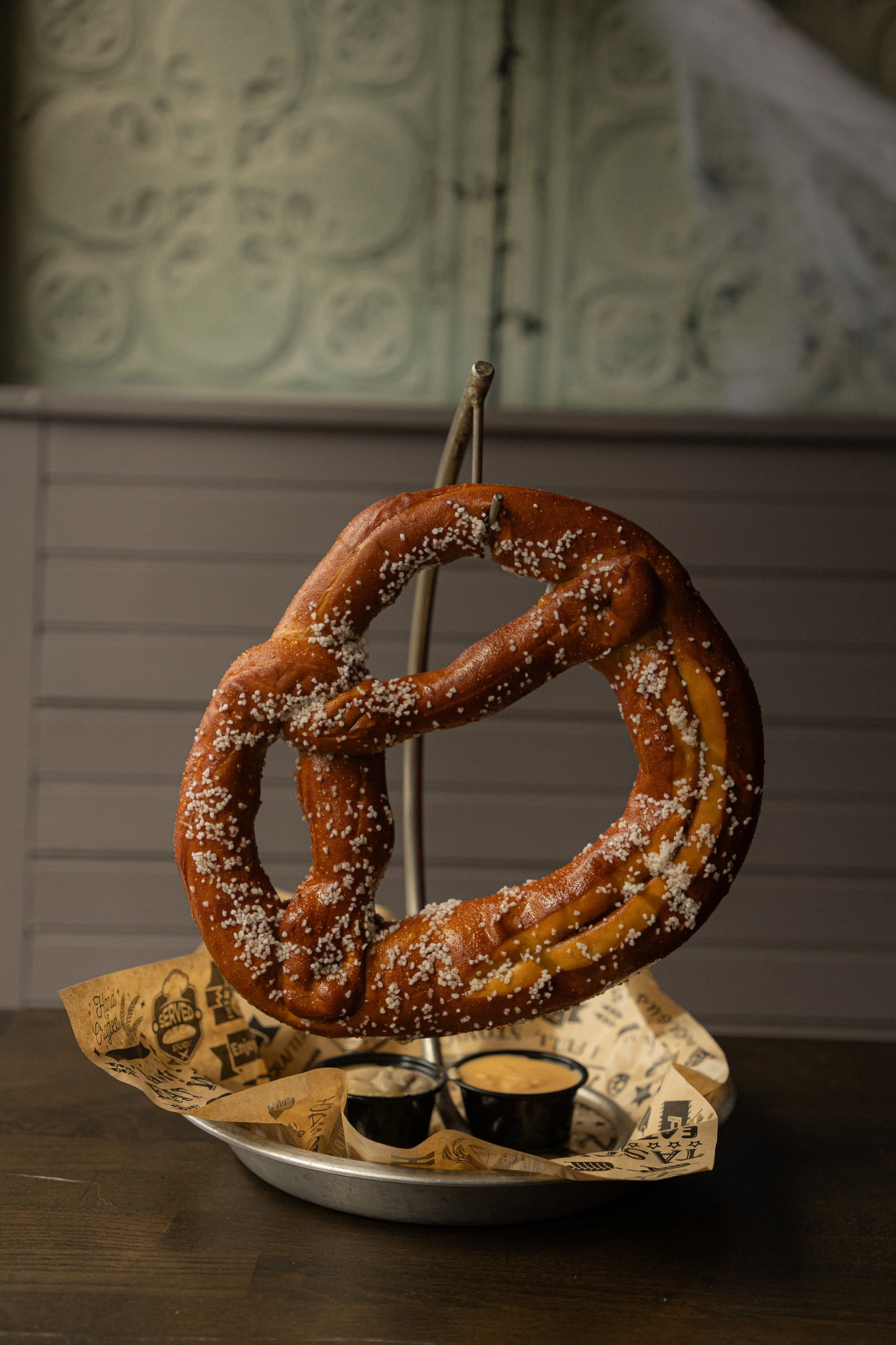 Hanging from a hook, a big pretzel covered in salt. Beneath it are two cups one with dijon mustard, the other a cheese sauce.