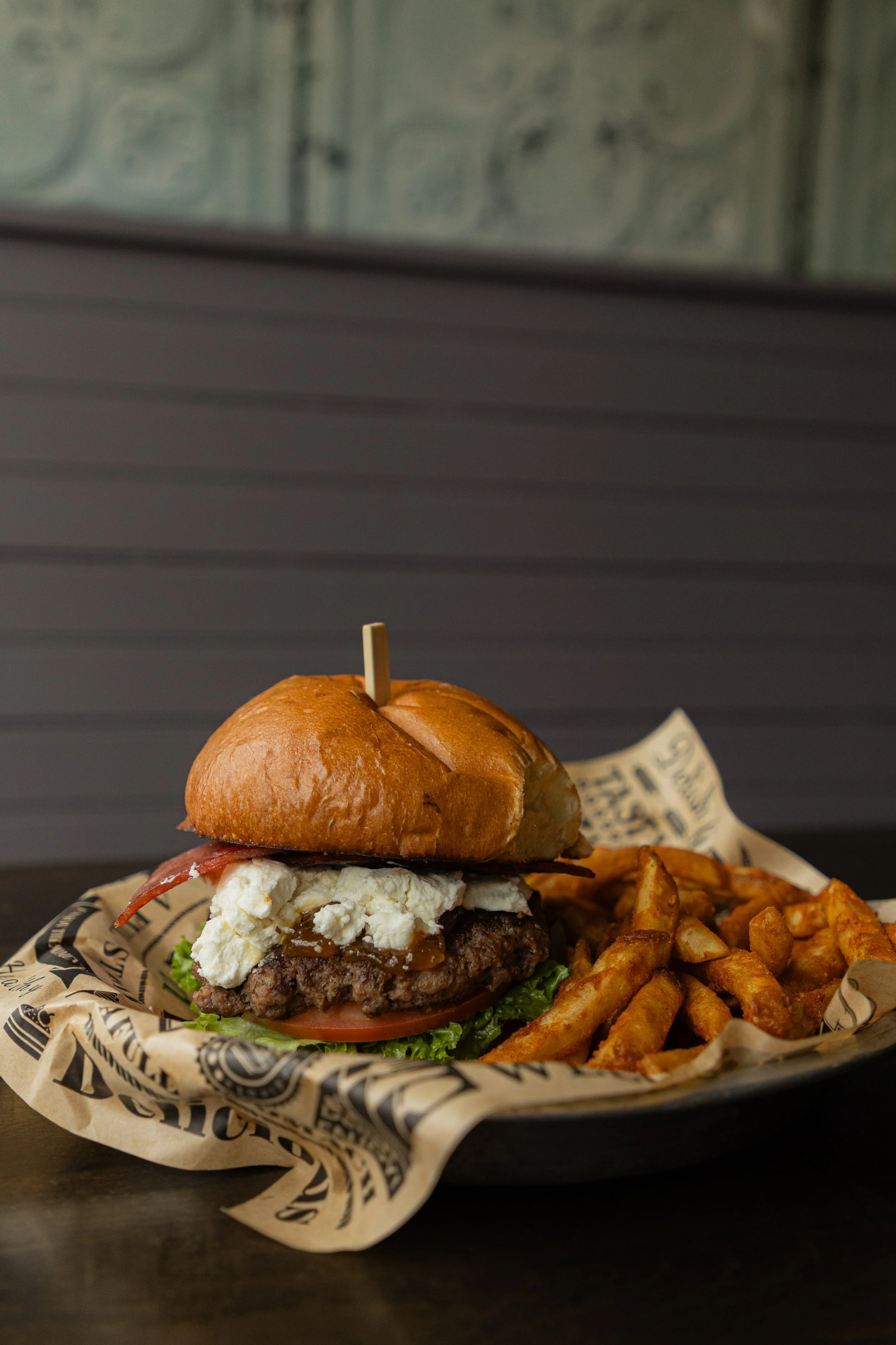 A burger on a bed of tomato and lettuce has prosciutto, fig jam & goat cheese spilling out the side.