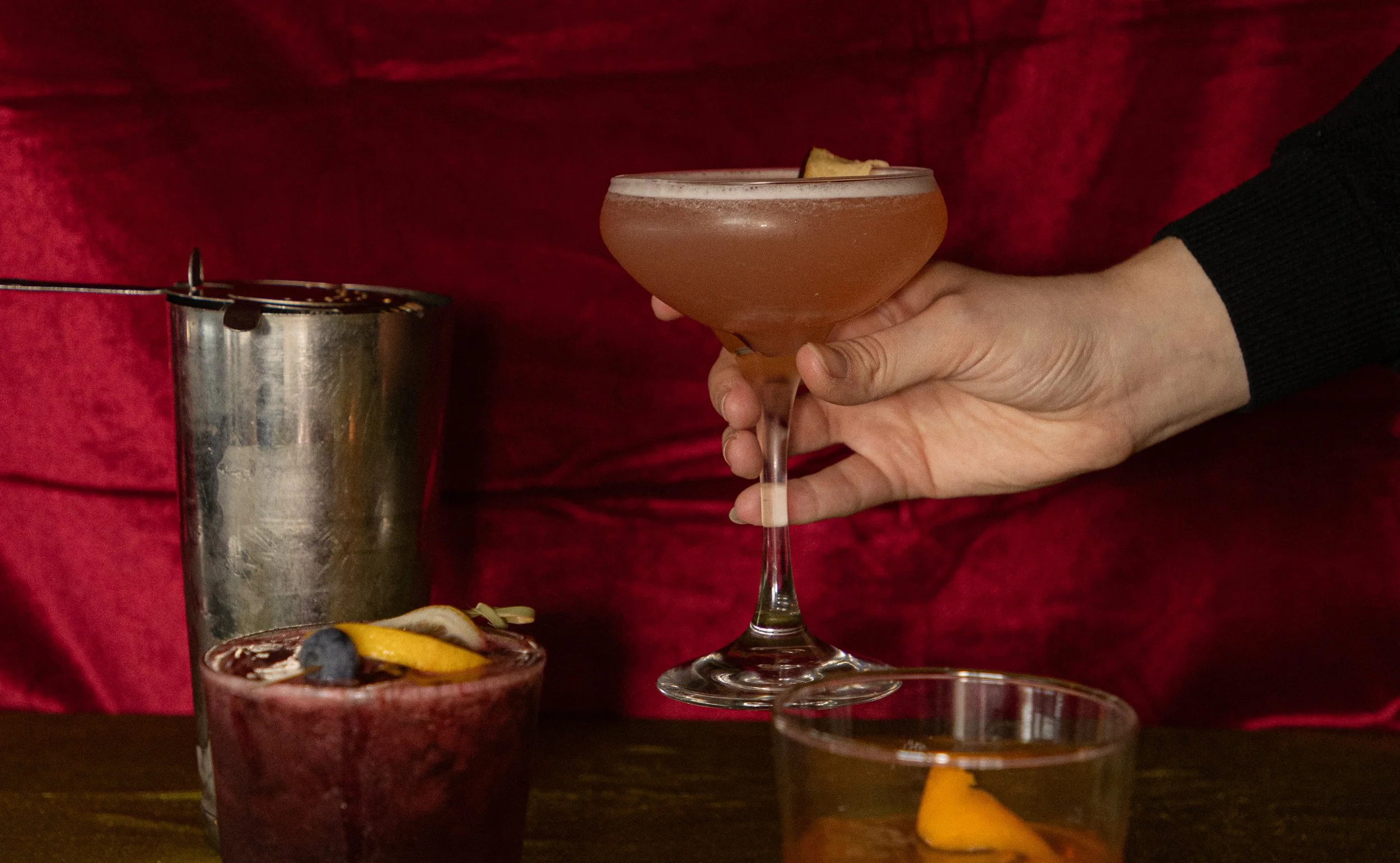 A hand brings in a cocktail with a red velvet backdrop.
