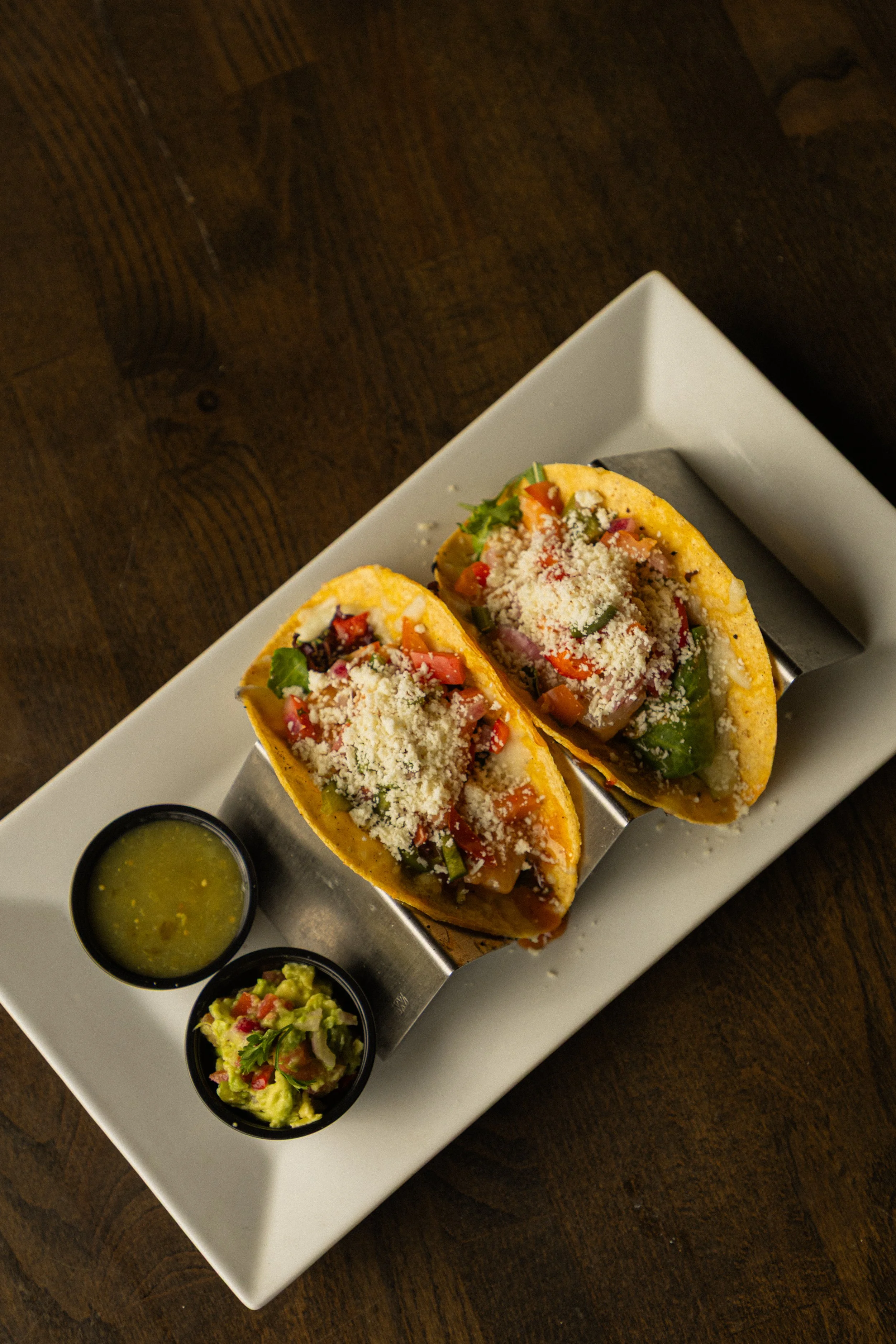 Overhead shot of two shrimp tacos topped with oaxaca cheese, tomatoes, lettuce. To the side is a cup of salsa verde and guacamole.
