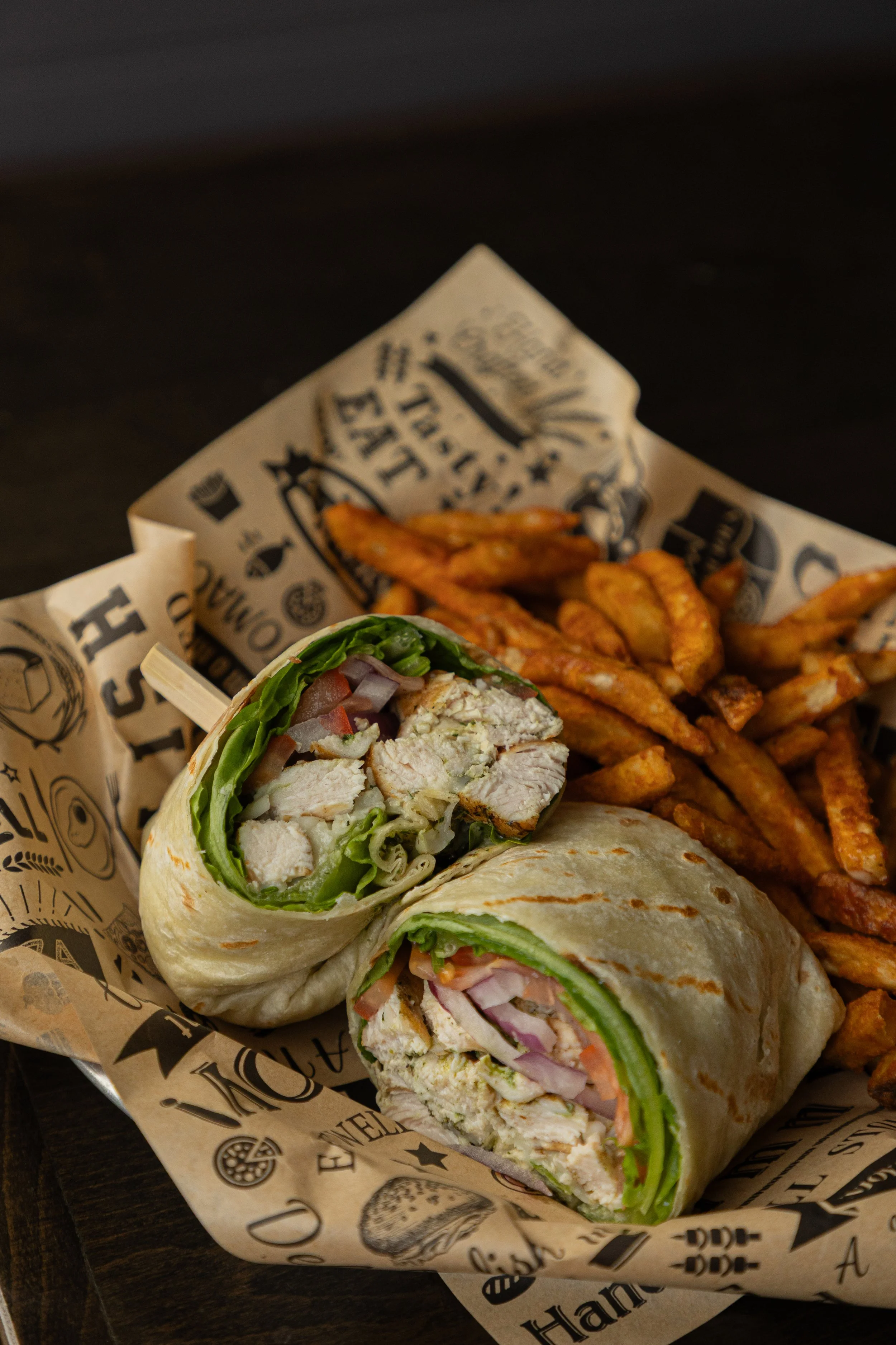 A chicken wrap cut open, inside there is red onions, tomatoes, lettuce, and grilled chicken. Behind it is a bed of french fries.