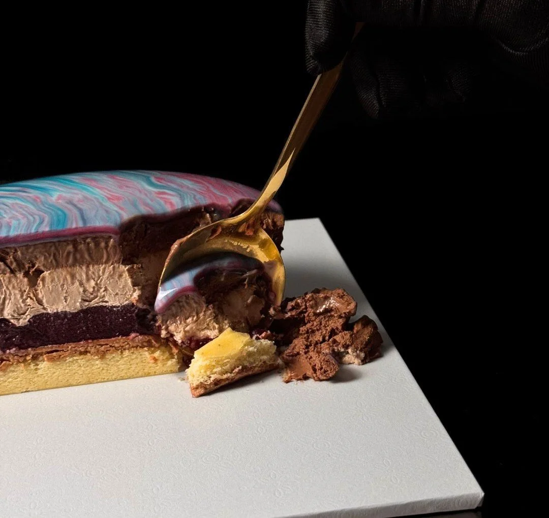 Slice of luxury entremet dessert by AFTERSIX, showing layered chocolate mousse with a marbled mirror glaze, served on a white base with a gold spoon.