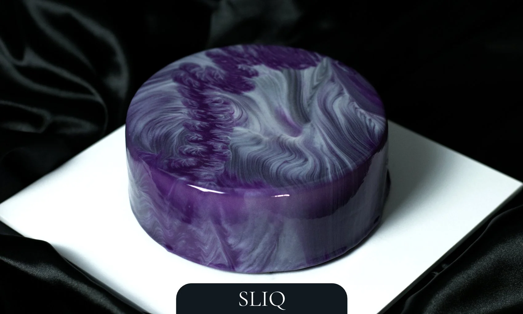 Luxury round entremet dessert by AFTERSIX with purple and grey marble mirror glaze, displayed on a white platform against a dark backdrop, tagged with the name 'SLIQ'.
