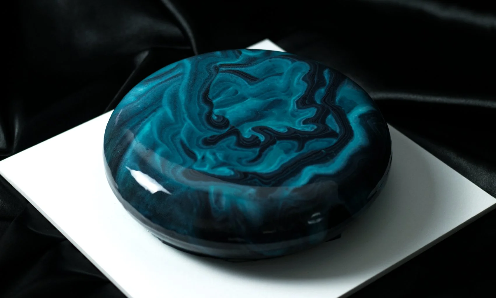 Luxury round entremet dessert by AFTERSIX with blue and black marble mirror glaze, placed on a white surface with a black fabric background, tagged with the name 'GRAND'.
