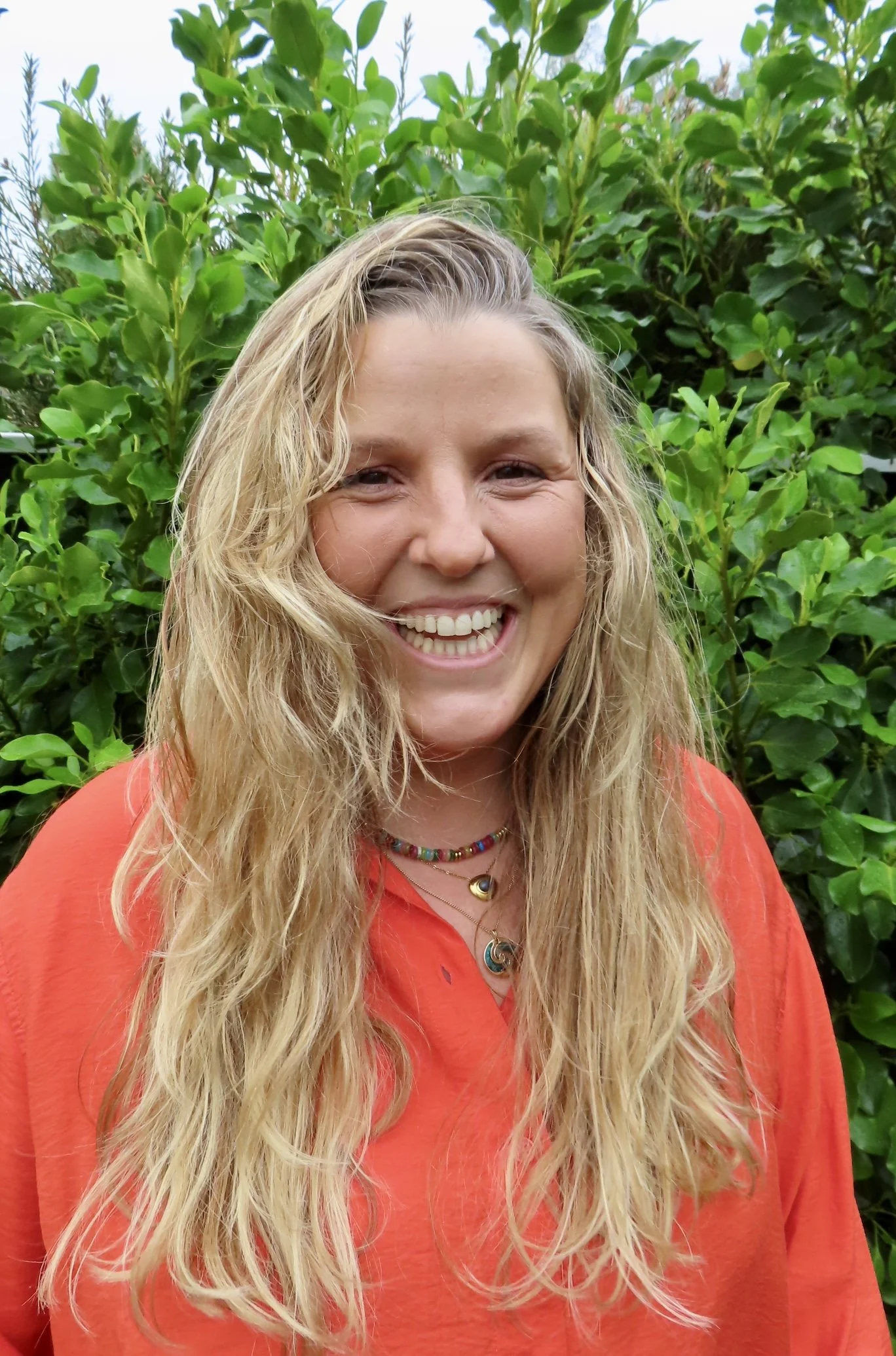 A smiling woman with long, wavy blonde hair standing outdoors in front of green foliage, wearing an orange shirt and colorful jewelry.