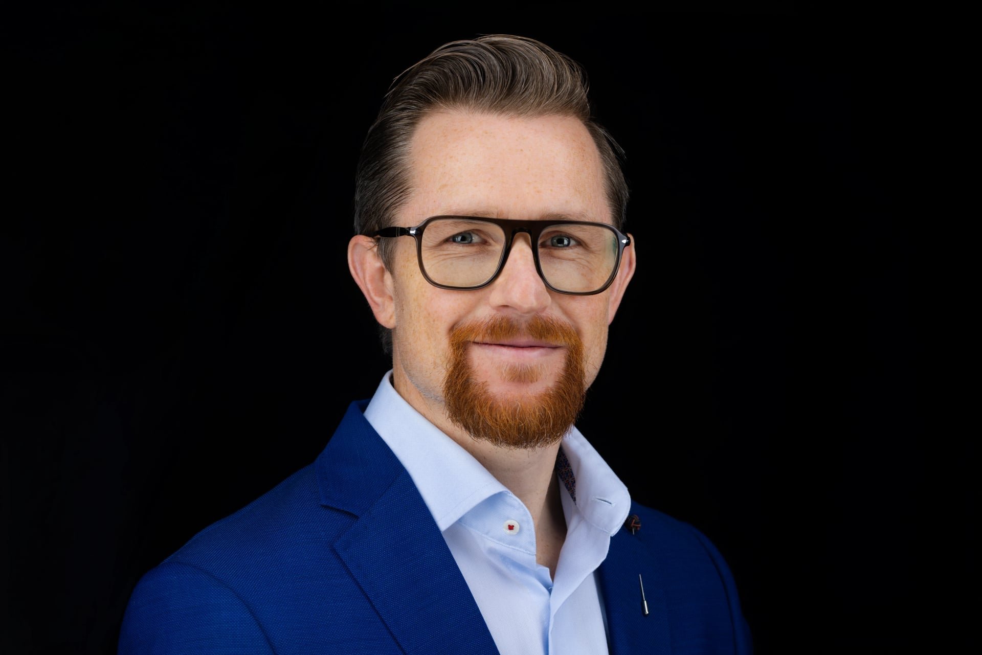 Portrait of a man with light skin, brown hair, glasses, a red beard and mustache, wearing a blue suit jacket and a light blue shirt, against a black background.