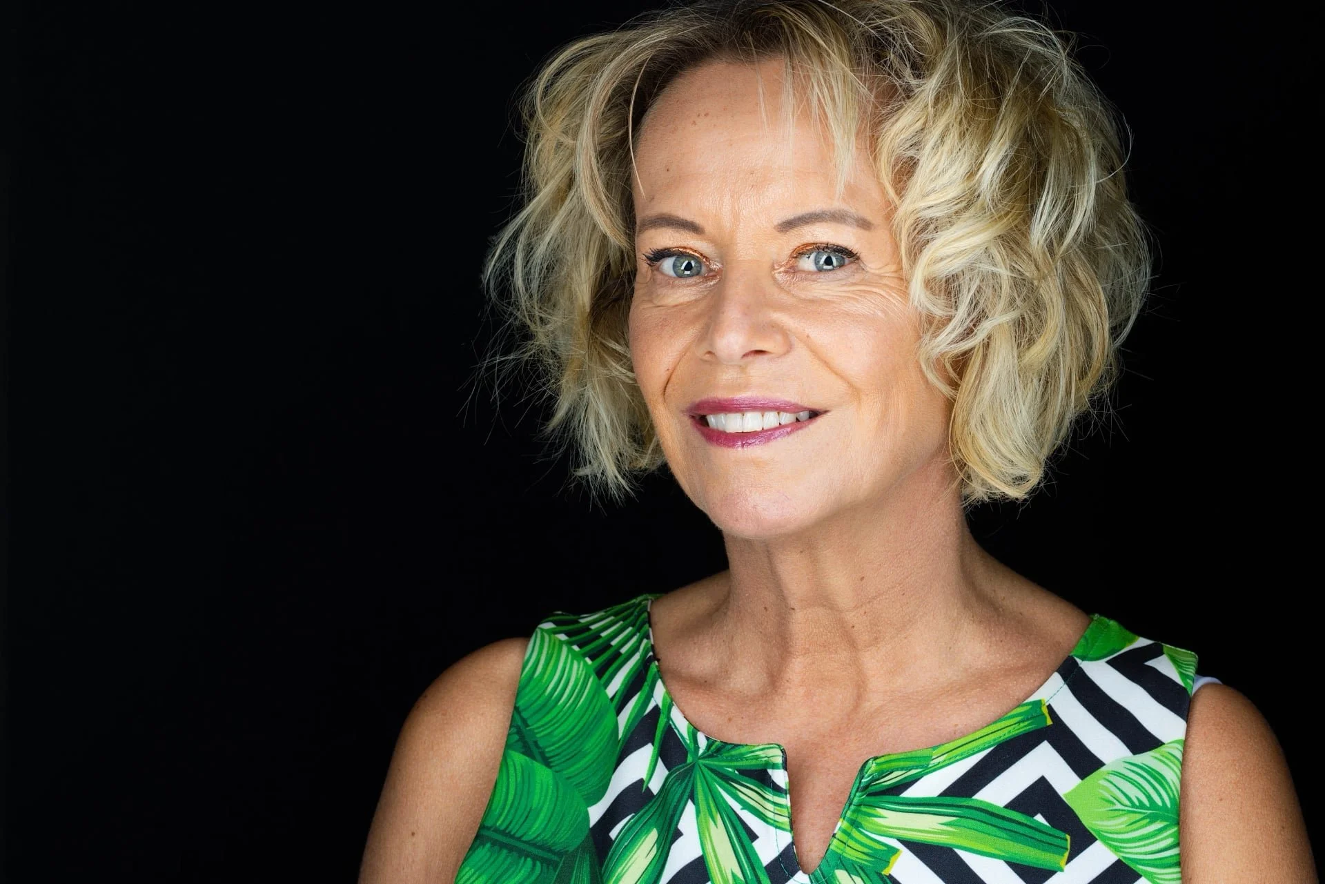 A smiling middle-aged woman with blonde curly hair, wearing a sleeveless dress with green tropical leaf and black and white geometric pattern, against a black background.