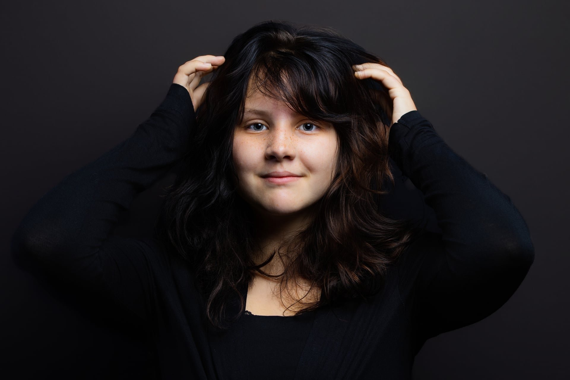 Portrait of a young woman with dark, wavy hair, blue eyes, and freckles, wearing a black top, posing against a dark background.
