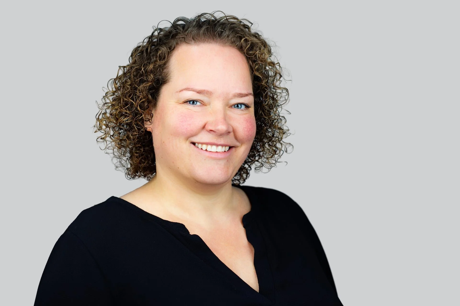 Headshot of a woman with curly brown hair, blue eyes, and a black top, smiling at the camera against a plain light gray background.