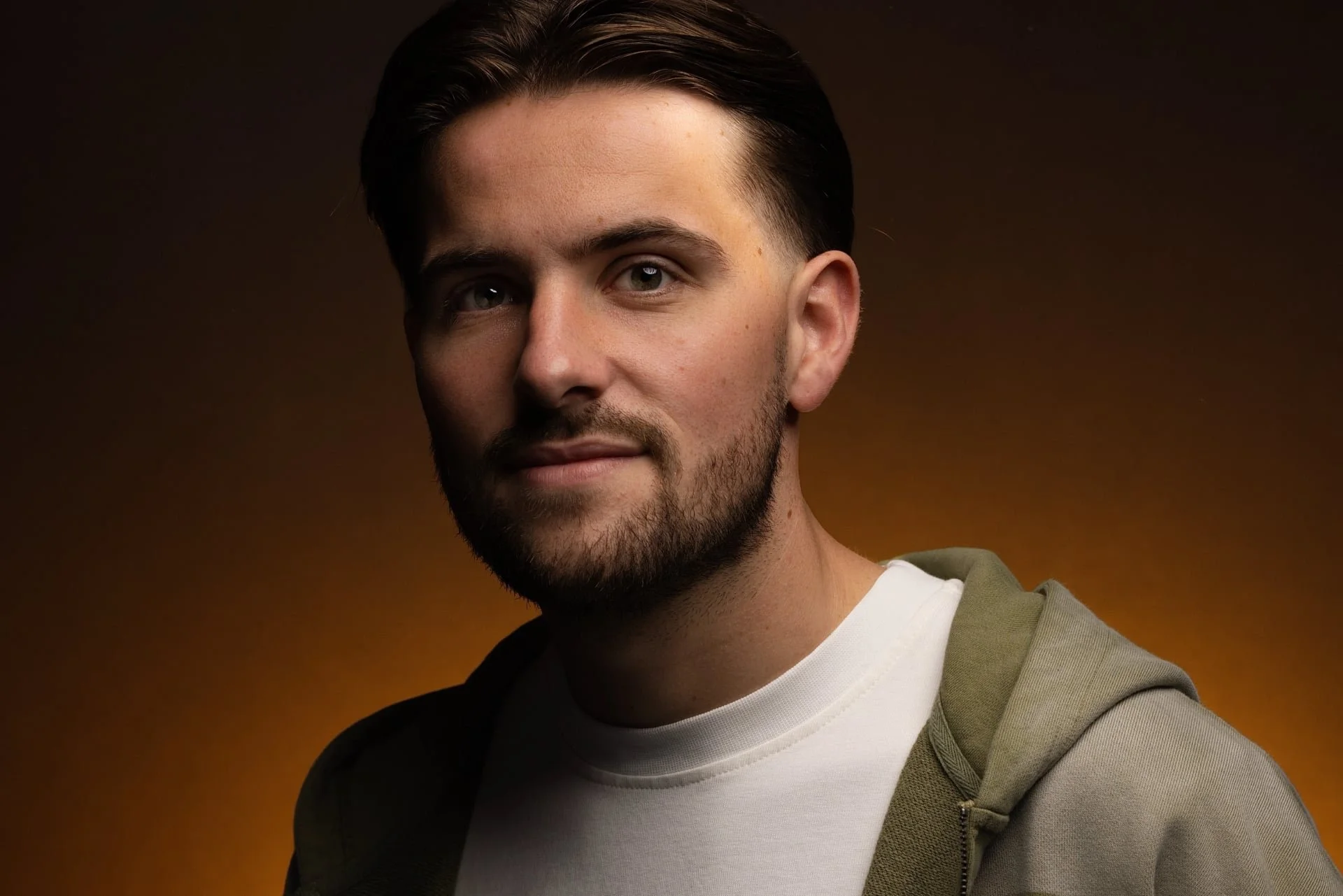 A young man with dark hair, a beard, and fair skin, wearing a white t-shirt and an olive-green hoodie, looking at the camera with a slight smile, against a brown gradient background.