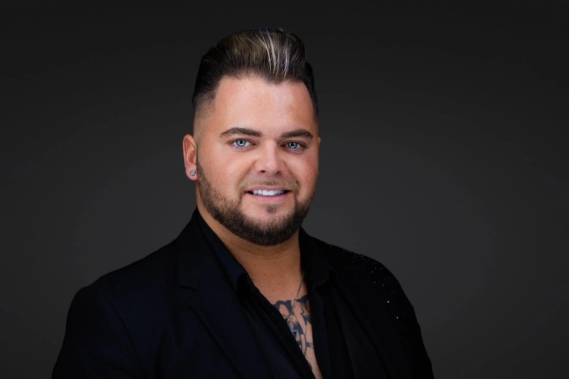 Headshot of a young man with blue eyes, dark hair with blonde highlights, a beard, wearing a black shirt and a small necklace, smiling against a dark background.
