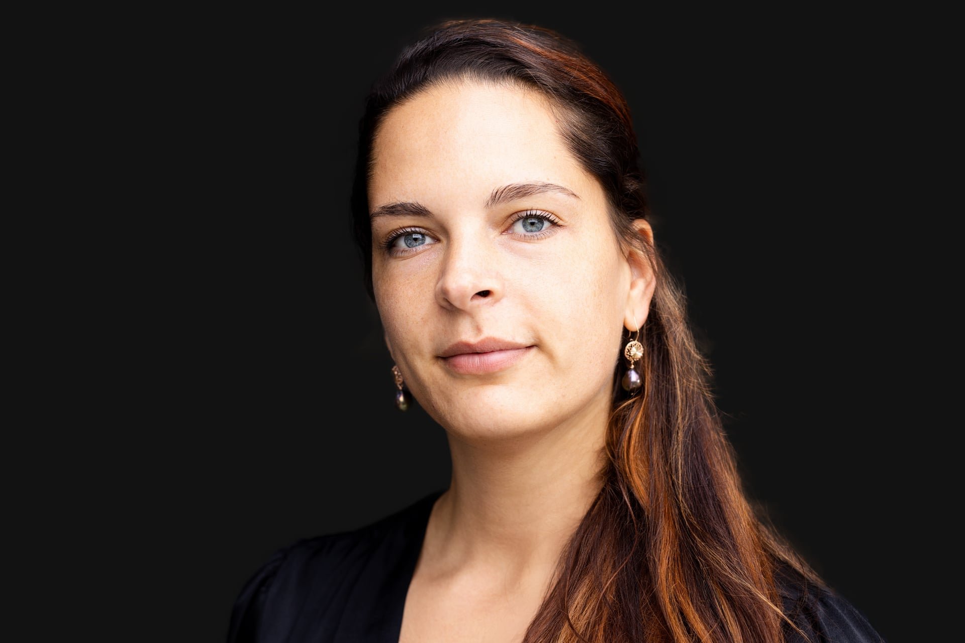 A woman with fair skin, blue eyes, and long brown hair with red highlights, wearing gold earrings with purple stones, against a dark background.