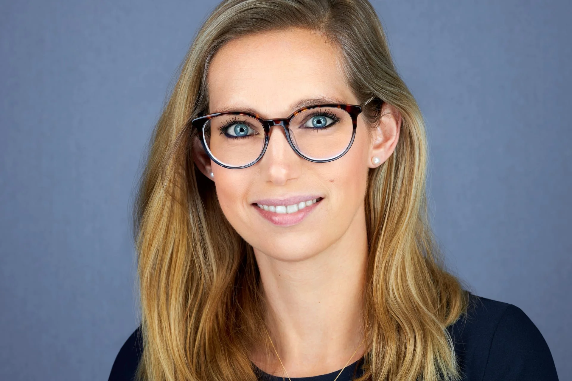 A woman with long blonde hair wearing glasses with a black and tortoise shell frame, small pearl earrings, and a black top, smiling against a plain, grayish-blue background.