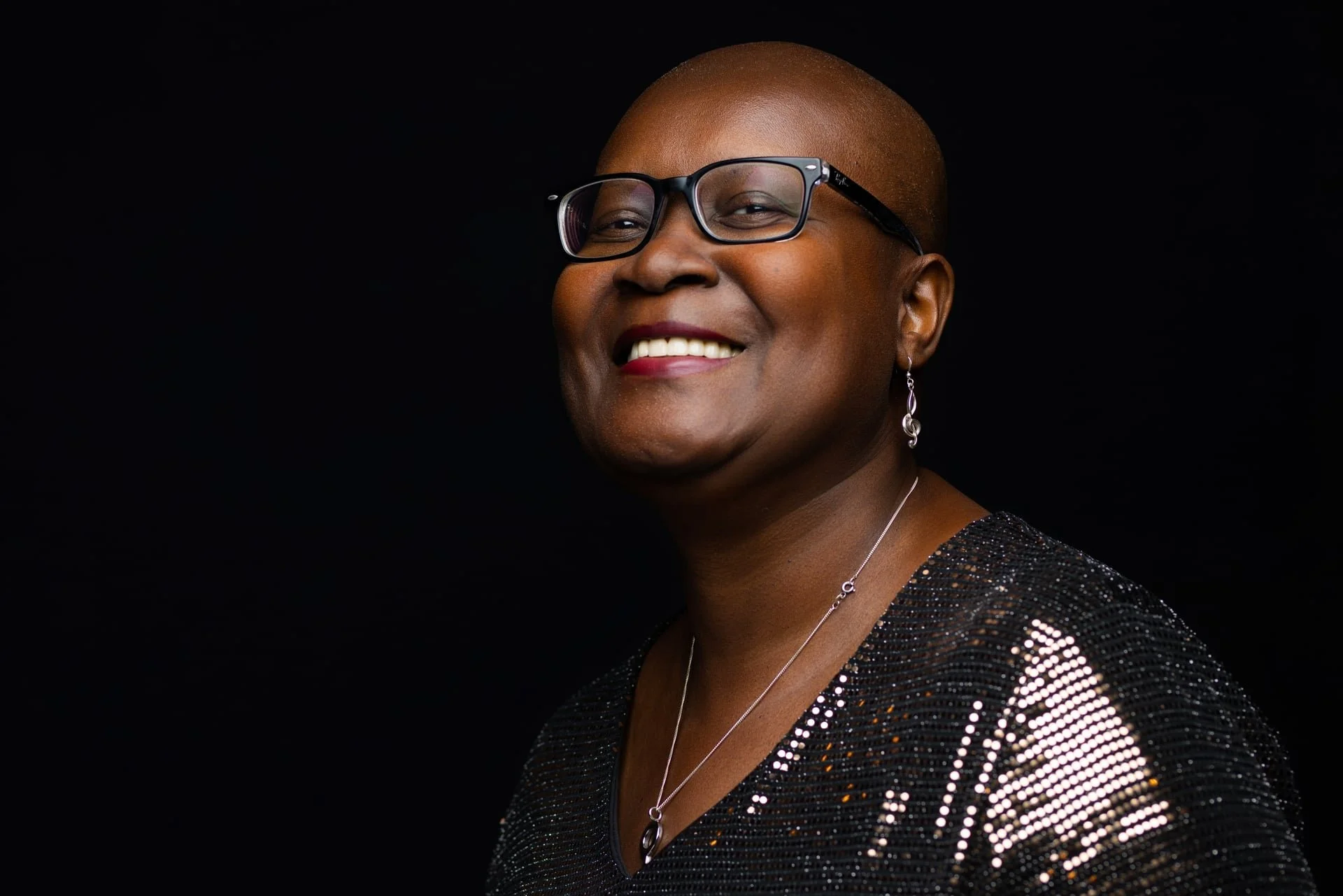Portrait of an African American woman with a bald head, wearing glasses, earrings, necklace, and a black sequin top, smiling against a black background.
