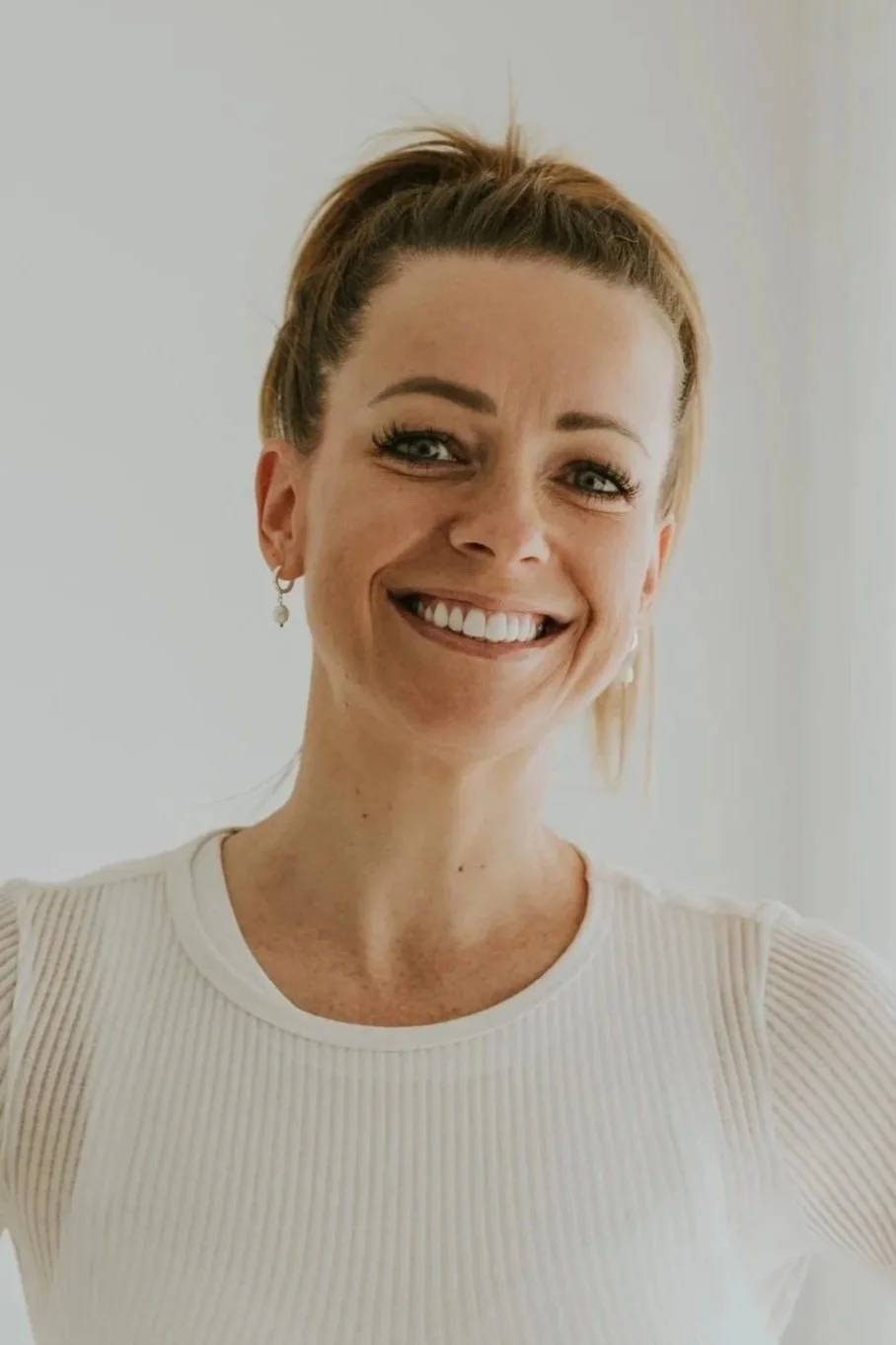 A smiling woman with blonde hair tied up, wearing earrings and a white ribbed top, posing against a plain background.