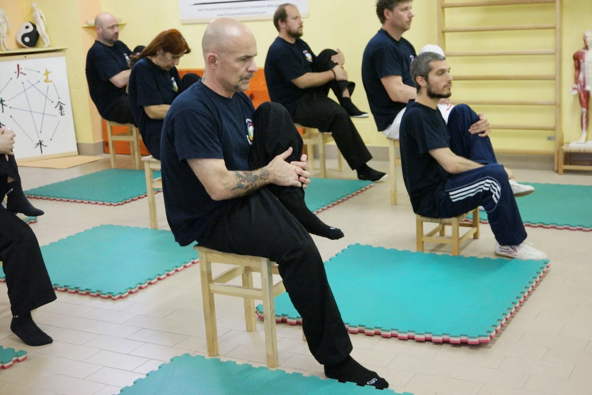 Gruppo di persone sedute su sedie, praticando esercizi di respirazione o meditazione, in una stanza con attrezzature per lo yoga o il Pilates.