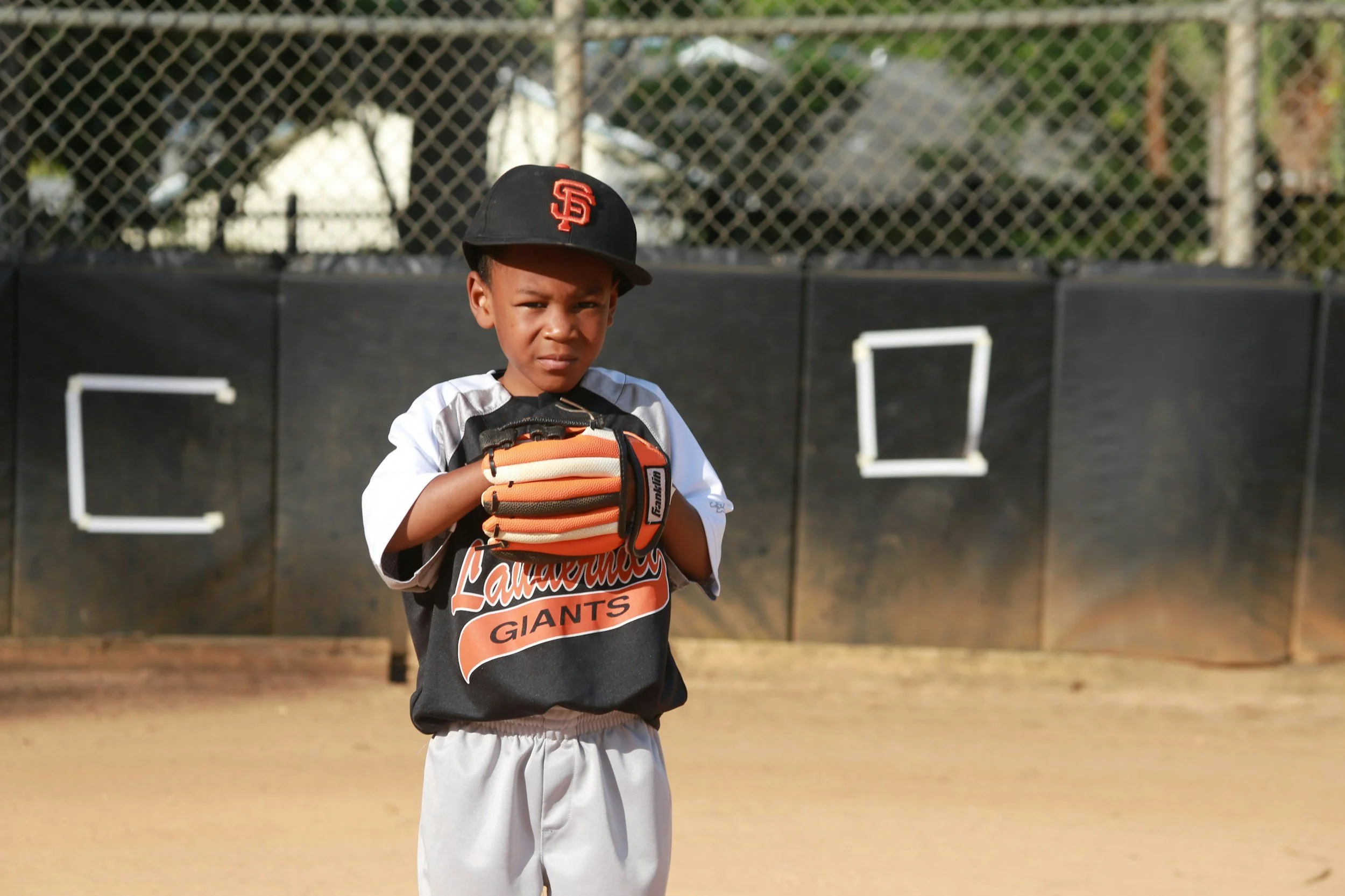 Bambino in uniforme da baseball con cappello nero e maglietta dei Giants, che indossa un guanto da baseball, in campo sul diamante.