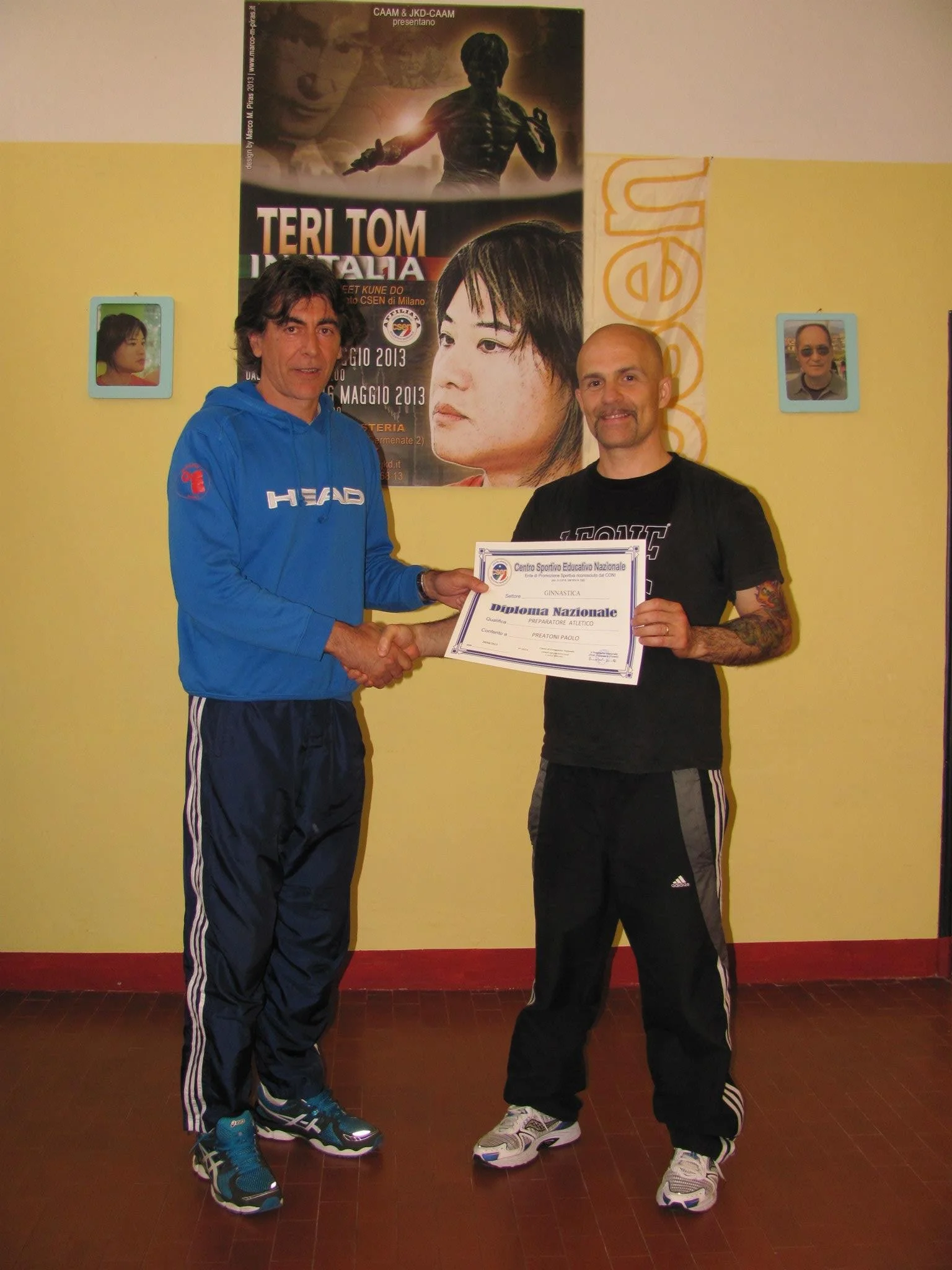 Due uomini che si stringono la mano mentre uno di loro riceve un diploma di riconoscimento, in un ambiente interno con poster e foto alle pareti.