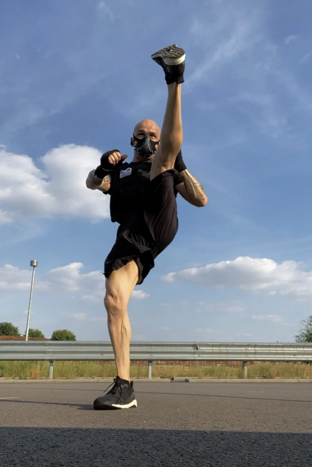 Uomo che esegue un calcio alto con la gamba destra, indossa una mascherina, abbigliamento sportivo nero, su strada asfaltata con cielo blu e nuvole leggere sullo sfondo.