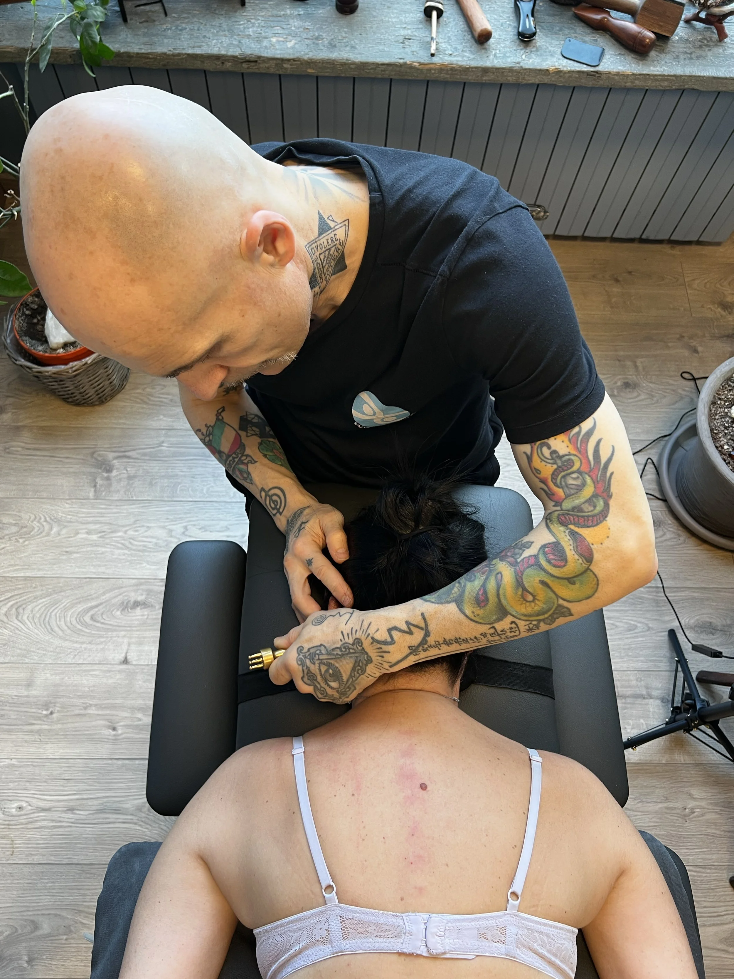 Tatuatore con braccia tatuate mentre fa un tatuaggio sulla schiena di una donna seduta su un tavolo in un ambiente di tatuaggi.