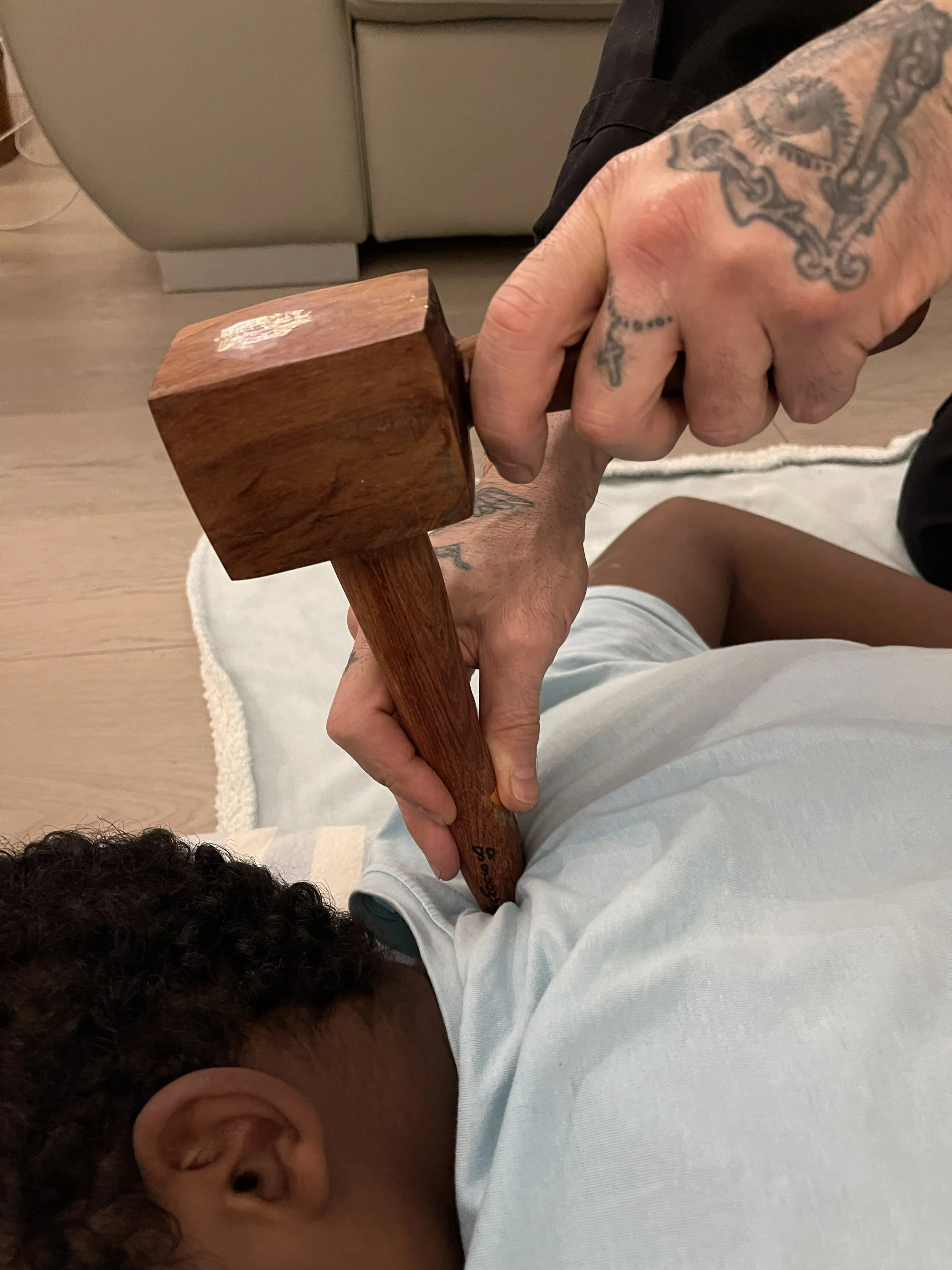 Persona con tatuaggi sta usando un martello di legno per picchiare un'altra persona che è sdraiata, con i capelli corti e ricci. La scena si svolge in un ambiente interno, con pavimento in legno e un divano grigio sullo sfondo.