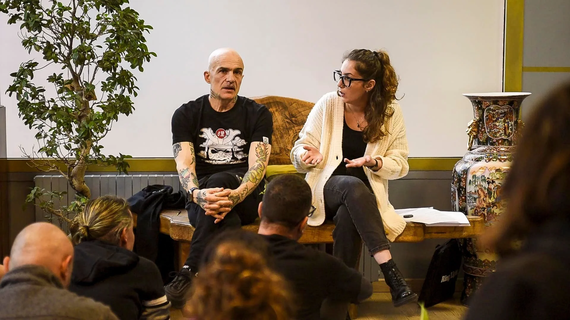 Una donna e un uomo seduti su una panca di legno che parlano con un pubblico seduto davanti a loro.