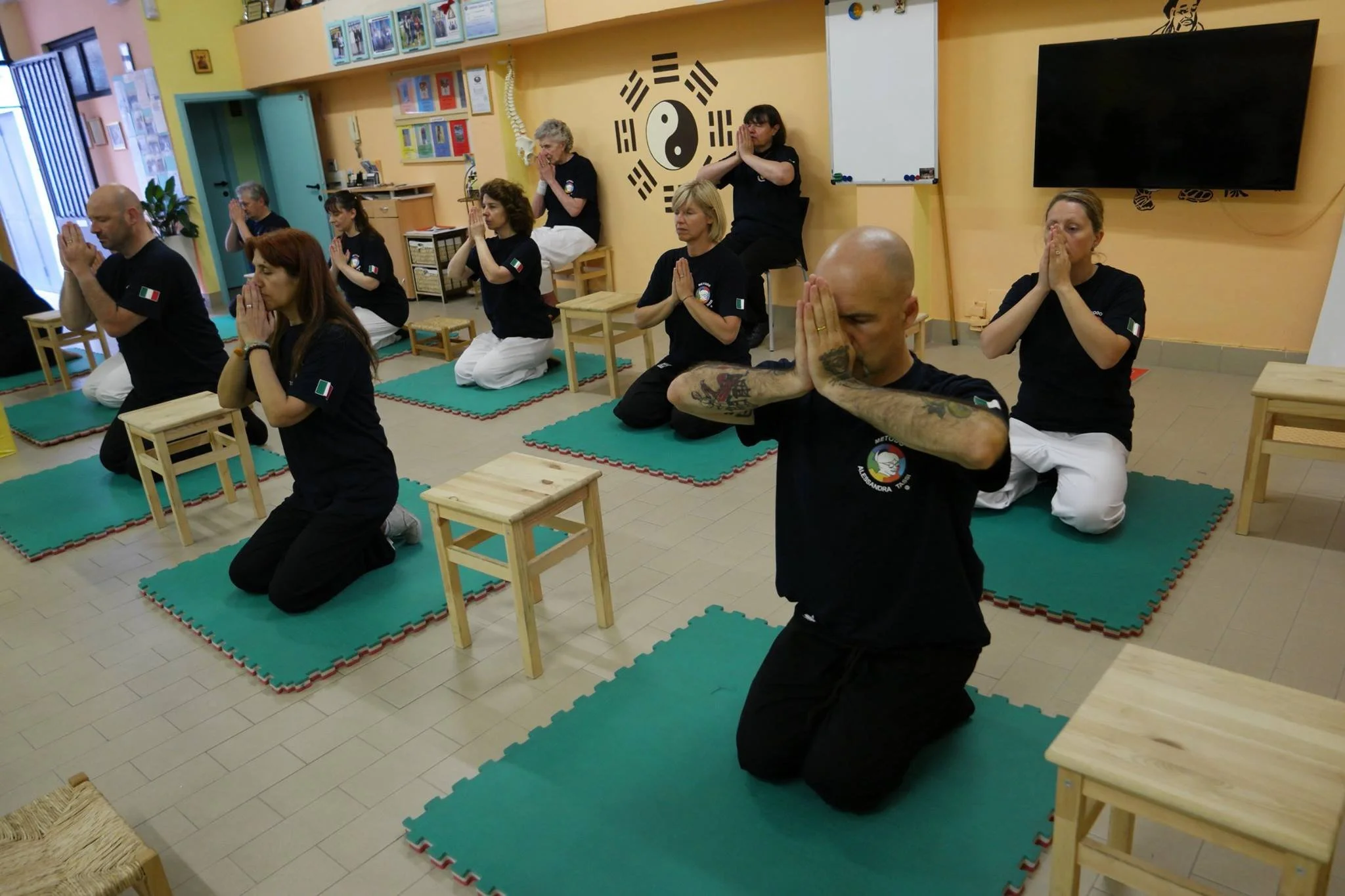 Gruppo di persone che praticano meditazione o mindfulness in una stanza con tappezzeria gialla, sedute a terra con ginocchia piegate, mani in preghiera, e un simbolo yin-yang sul muro.
