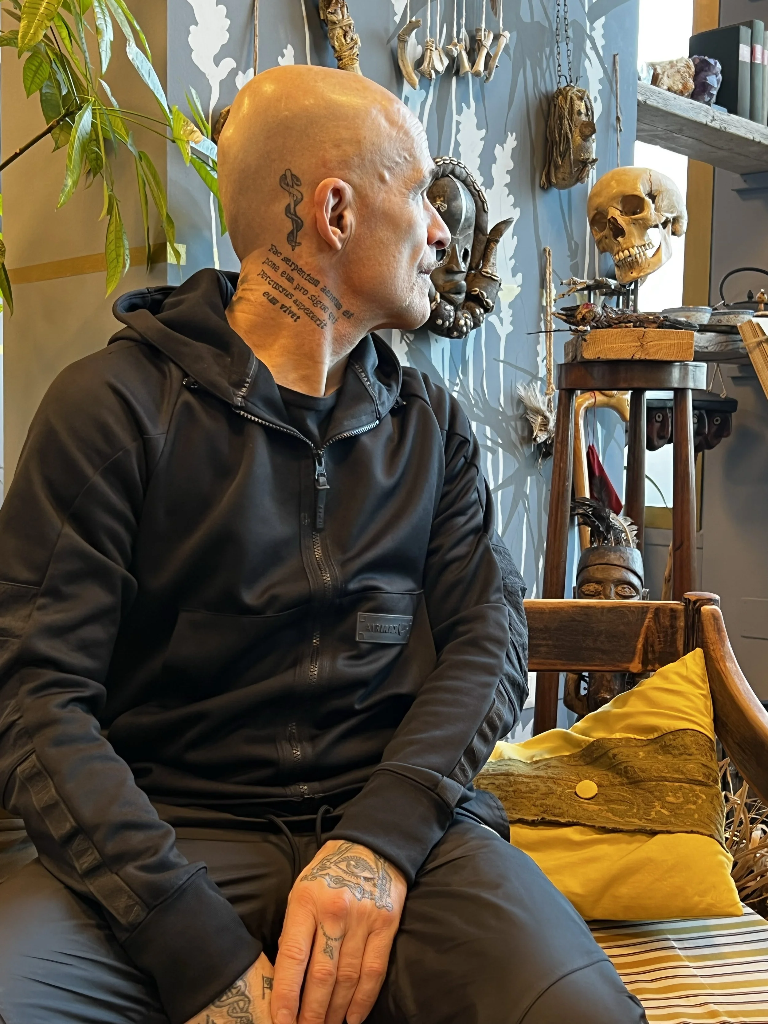 Uomo tatuato con cappuccio nero seduto in una stanza decorata con oggetti a tema macabro, tra cui teschi e maschere, in un ambiente con pareti decorate e scaffali.